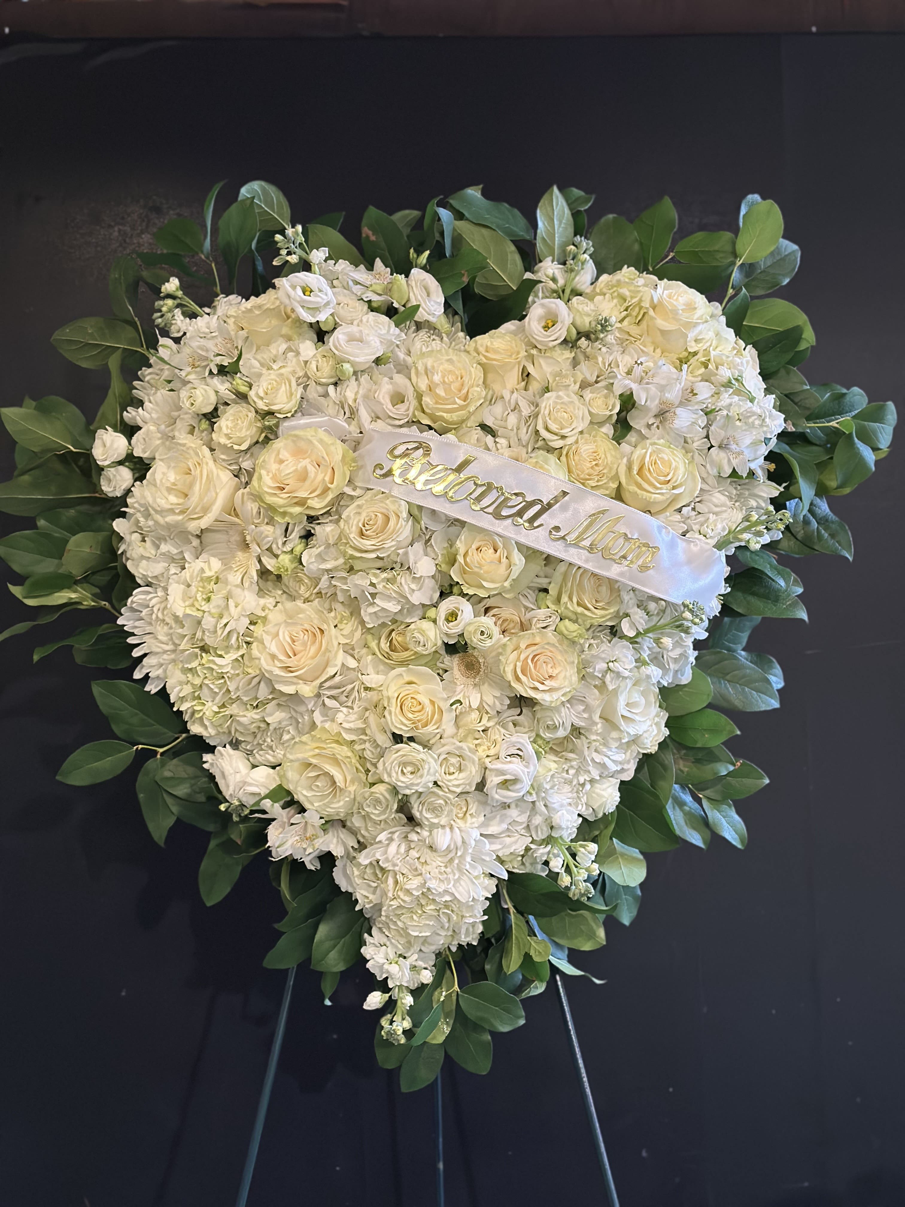 Heart-shaped white floral wreath with a ribbon reading "Beloved Mom"