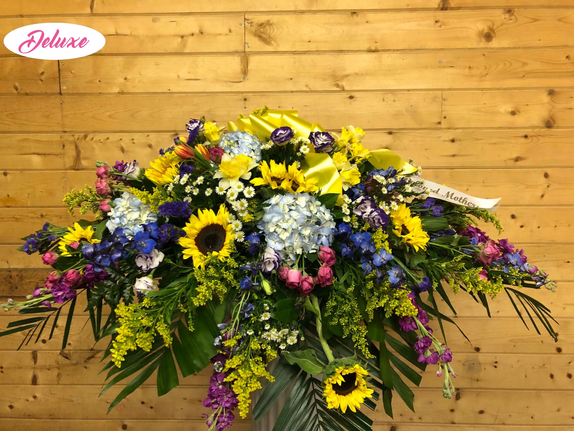Eternal Light - Honor a life beautifully lived with Eternal Light, a breathtaking casket cover designed to symbolize love, hope, and everlasting remembrance. This radiant tribute features lush blue hydrangeas, vibrant sunflowers, striking dark blue delphinium, hot pink spray roses, delicate goldenrod, and fragrant purple stock. Together, these blooms create a meaningful blend of warmth, serenity, and vibrant beauty.  Handcrafted by our expert florists, Eternal Light offers a graceful expression of love and remembrance, making it a heartfelt centerpiece for funeral or memorial services. Each arrangement is thoughtfully designed with the freshest flowers available, ensuring a beautiful and lasting tribute that celebrates a cherished life with dignity and compassion.