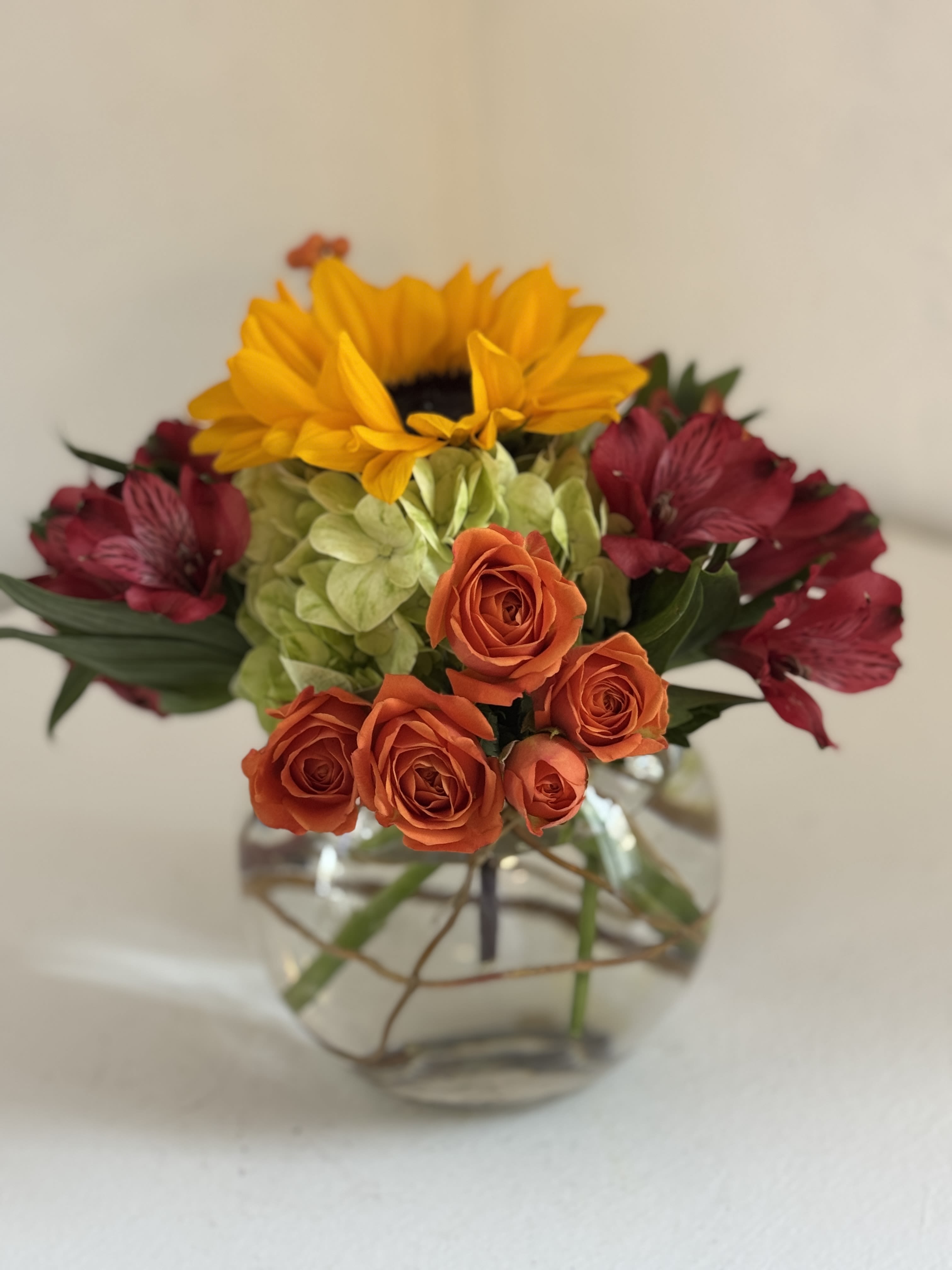 Mixed bouquet with orange roses, yellow sunflower, green hydrangea, and burgundy flowers in a glass vase