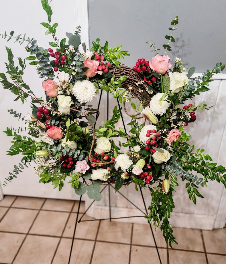 Wintery Wreath - A lush grapevine wreath arranged on a black metal easel, this large wintery wreath blends classic holiday charm with a calm, reflective mood perfect for holiday, sympathy and funeral, or winter displays. Clusters of cream and blush roses, white cushion chrysanthemums, and open and budded lilies are artfully spaced around the natural vine ring so the rustic base still shows through; bright red berry stems and abundant greenery add seasonal color and texture that also work nicely for Hanukkah, Memorial Day, or a housewarming moment.  The design features mixed reds, pinks, purples, lavenders, whites, and greens for a layered, garden-fresh look that feels both elegant and natural. Blooms and foliage cascade slightly at the bottom for movement and softness, giving the wreath visual depth from every angle. Delivered ready to display on the included metal easel, it's a thoughtful way to say "I'm thinking of you," "thank you," "I'm sorry," or simply "just because" - a wreath that brings comfort, seasonal cheer, and a welcoming presence to porches, entryways, or memorial settings.