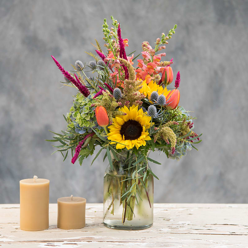 Summerly - Bright summer colors and happy blooms including sunflowers, tulips, snapdragons and thistle in a unique milk jug vase.