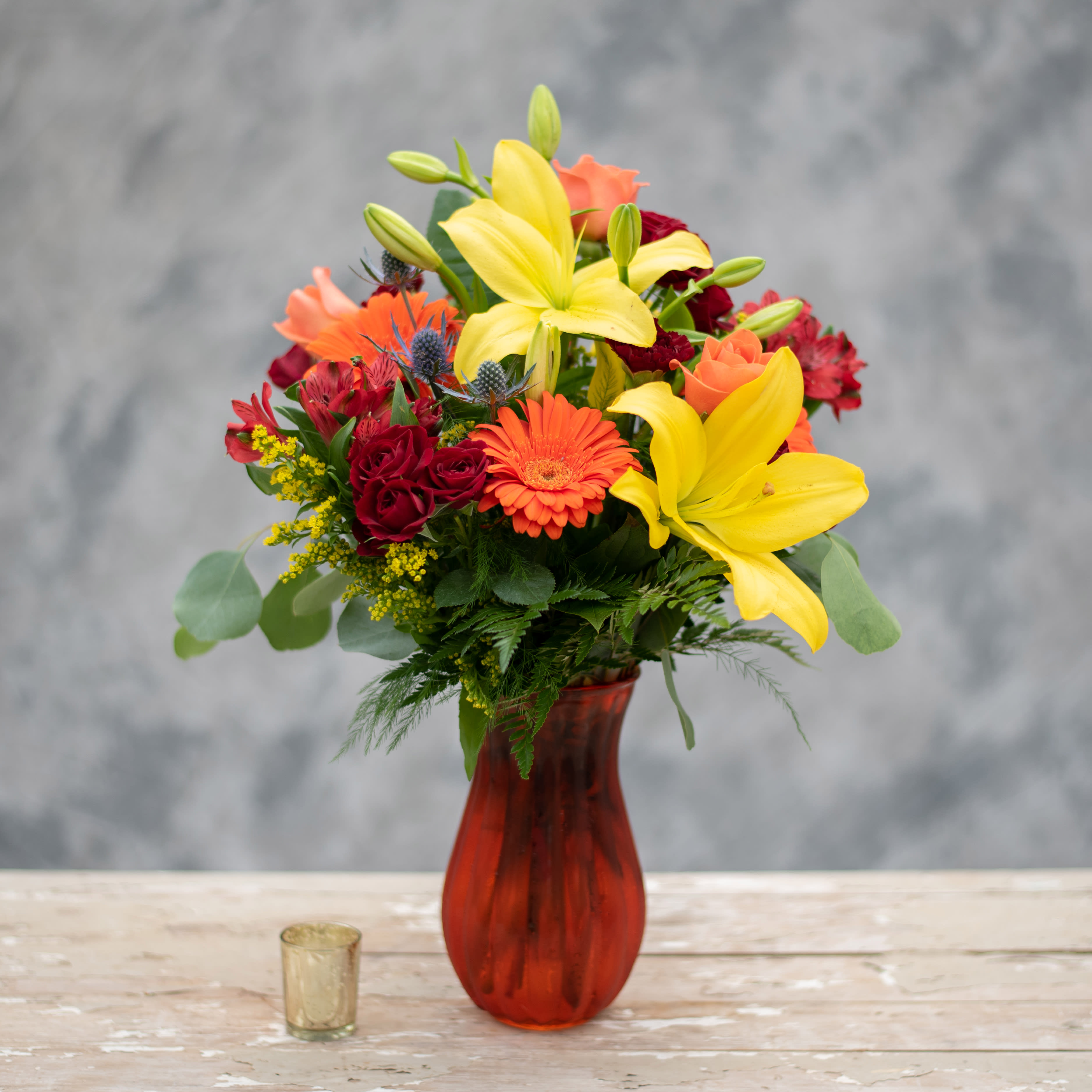 Mixed bouquet with yellow lilies, orange gerbera daisies, and red roses in a red vase