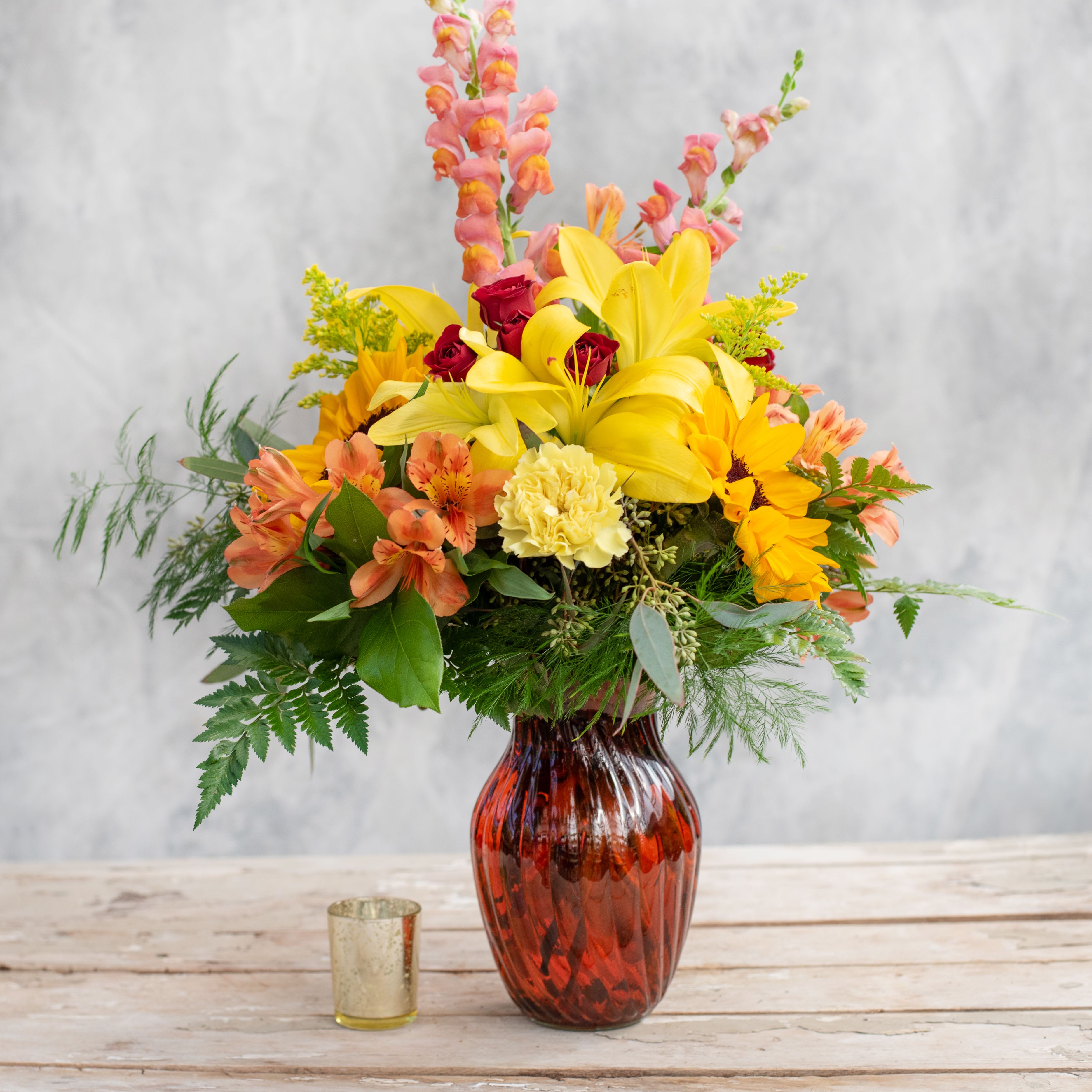 Amberly - Bright yellow sunflowers, red spray roses, snap dragons and lilies among some unique fall greenery and purple status all nestled in a vibrant fall vase. Stunning!