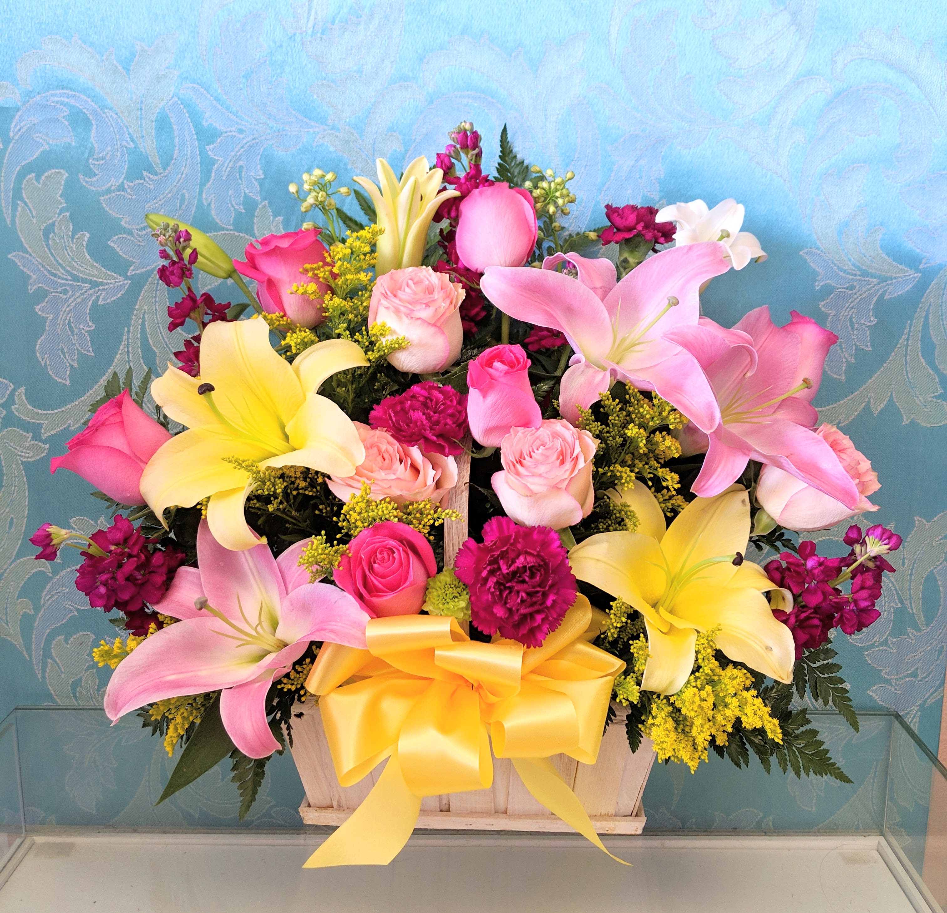 Pink and yellow lilies with roses in a wooden basket tied with a yellow bow