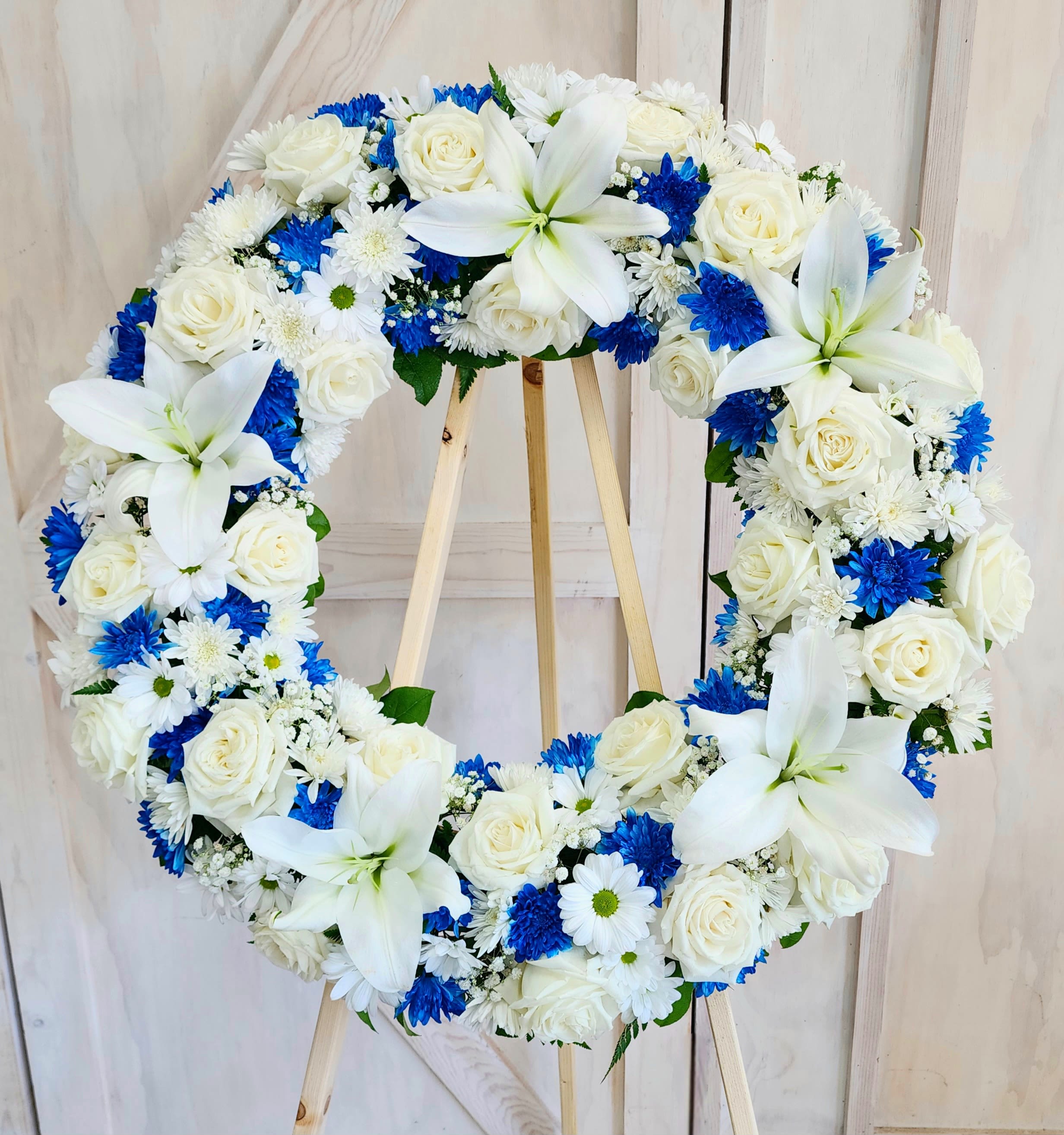Heart-shaped floral wreath with white roses, lilies, and blue daisies on a wooden stand