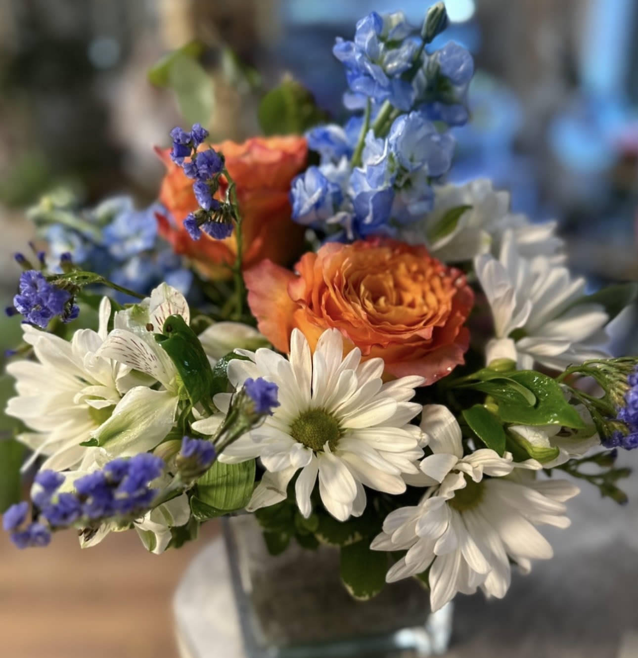 Bouquet of white daisies, orange roses, and blue flowers in a glass vase
