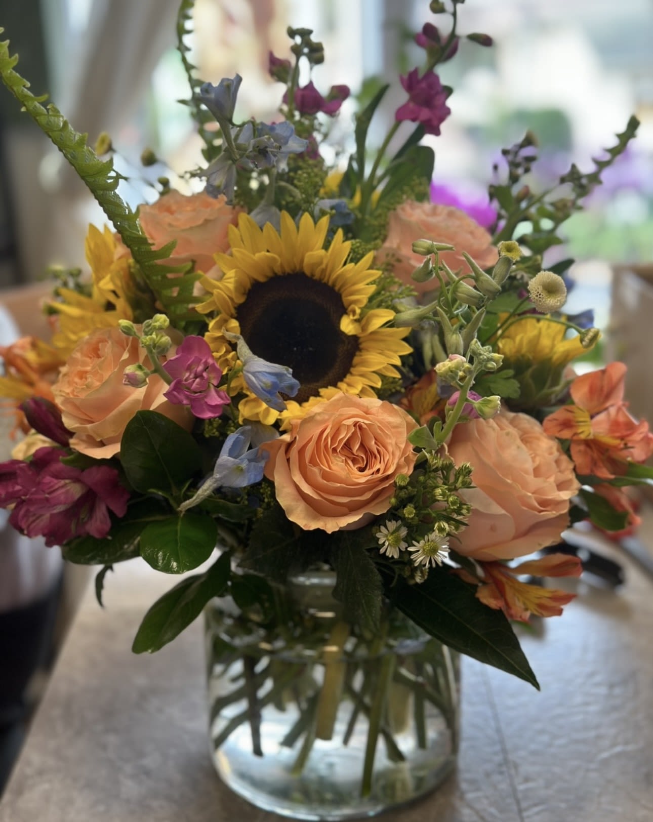 Mixed bouquet with a sunflower, peach roses, and purple blooms in a glass vase