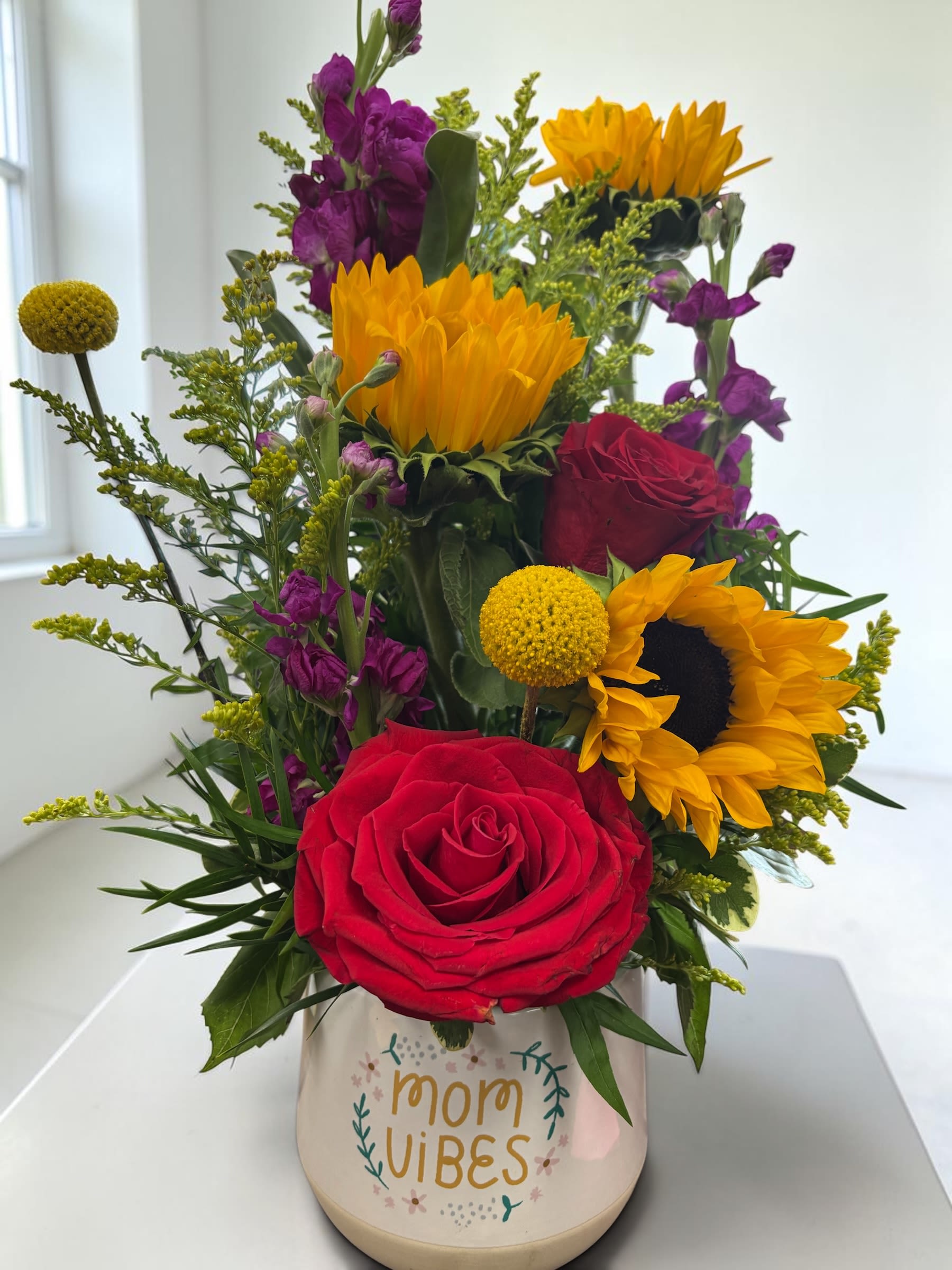 Mom vibes - Bright yellow sunflowers paired with deep red roses and pops of purple create an immediate burst of warmth and playfulness-an ideal flower arrangement for Mother's Day or to celebrate a special mom any day. This upright "mom vibes" bouquet reads as cheerful and celebratory, the kind of gift that makes the person you're thinking of feel seen and cherished the moment it arrives.  Hand-arranged in a cream ceramic container printed with "mom vibes," the design mixes saturated warm and cool tones for a full, lively look that brightens a kitchen table or bedside. The arrangement combines classic roses, bold sunflowers, and complementary purple accents for contrast and texture, presented compactly for easy display.  Perfect for moms who love color and personality, this composition sends a joyful, affectionate message-whether for Mother's Day, a birthday, or a simple thank-you. The ceramic vessel is ready to set out as-is; swap the water every couple of days and trim stems slightly to extend freshness.