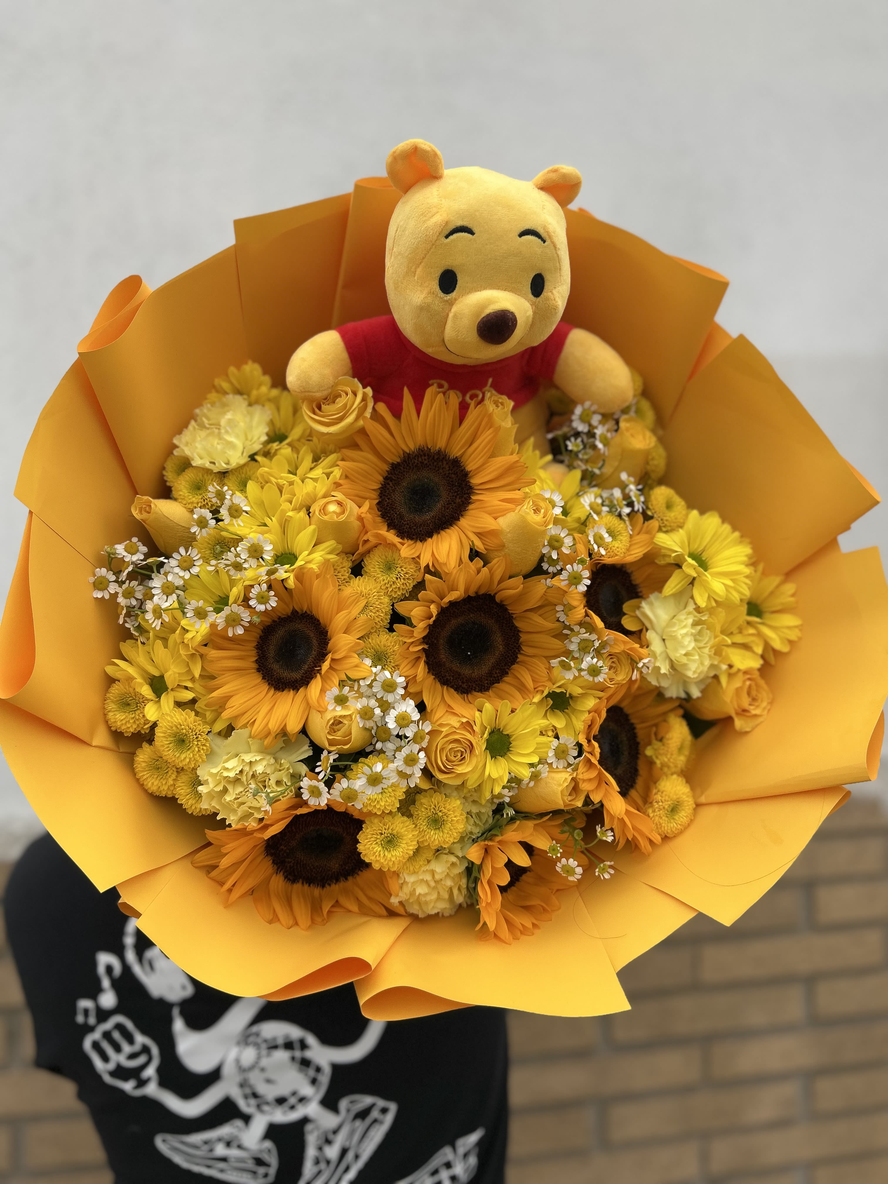 Oh Sunny Day Yellow Rose and Sunflower Arrangement - Sun-soaked yellow roses, cheerful sunflowers, and dainty white daisies combine in a hand-tied flower arrangement that reads like a warm hug - the perfect "just because" surprise or a bright token of love and romance. Oh Sunny Day is a playful anniversary or everyday bouquet designed to make the person you're thinking of feel seen and celebrated; it's also a joyful choice for Valentine's Day, Mother's Day, or a quinceañera celebration in Hawthorne, CA.  This full, rounded bouquet layers large golden sunflowers and yellow roses with soft yellow carnations, button-style chrysanthemums, and tiny white daisies for rich texture and sunny contrast. An orange-yellow paper wrap fans out around the stems to amplify the warm palette, and a small plush bear tucked among the blooms adds a whimsical, gift-ready touch. The arrangement's size and dense composition create a substantial presence on arrival - the kind of bouquet that brightens a room and keeps smiling.  Send this arrangement when you want to lift someone's spirits or mark a loving milestone: she'll feel genuinely cheered by the radiant colors and the thoughtful, playful details. For care, keep the stems in fresh water and trim a half inch every few days to extend vase life; order same-day delivery in Hawthorne, CA when available to ensure it arrives at peak freshness.
