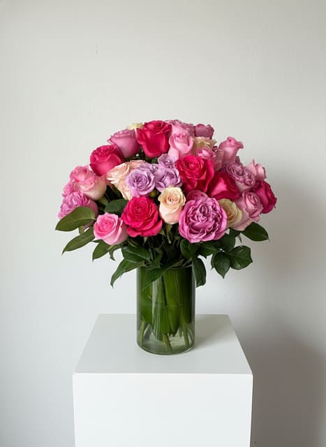 And i Love Her - A lush dome of roses in hot pink, blush, lavender and pale cream greets the eye-perfect as a birthday bouquet, anniversary roses arrangement, or a just-because gift to brighten someone's day. The rounded, tightly packed shape creates an immediate impression of abundance and romance, so the person you're thinking of will feel truly seen the moment this arrives.  Carefully gathered into a clear cylindrical glass vase with glossy green foliage at the base, the compact arrangement reads polished and classic on any table or bedside. The mix of warm pinks, cool lavender and soft white offers a feminine, versatile palette that complements many décor styles; its neat dimensions make it easy to display in small spaces. Order with confidence for celebrations or comforting gestures-simple water changes and fresh cuts at delivery will keep the roses looking their best.