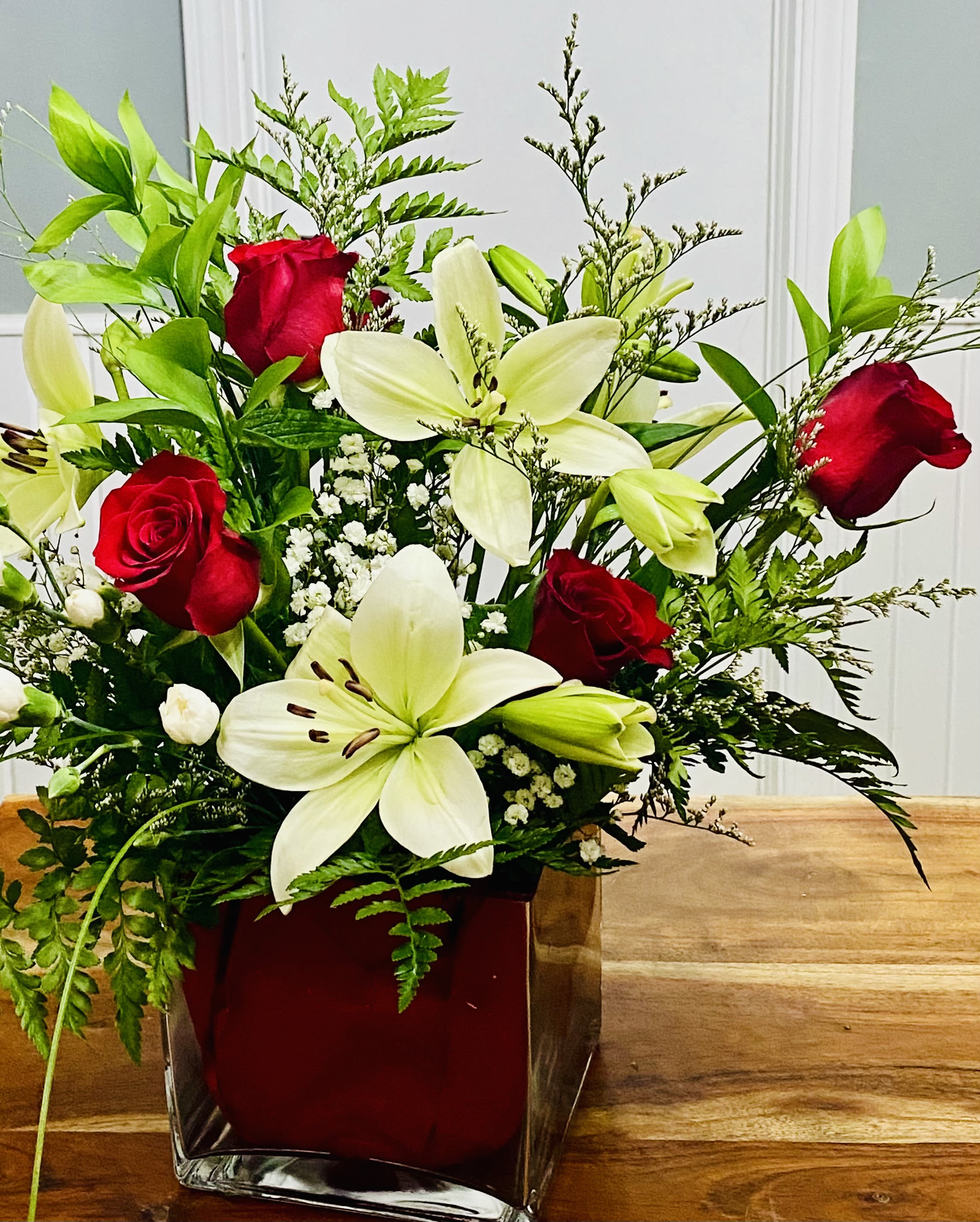 Heart of the Lily  - Open white lilies paired with deep red roses create an unforgettable contrast in this flower arrangement, perfect as a romantic anniversary bouquet or a heartfelt gift for a birthday, get well, or "I'm sorry" moment. Heart of the Lily reads as affection and encouragement: the luminous white lilies and red roses send a message of love and warmth that makes the person you're thinking of feel truly seen.  This mixed bouquet includes creamy white lilies, red roses, white carnations, and delicate white filler blooms arranged with lush foliage and ferns for a slightly dramatic, vertical silhouette. The blooms are gathered in your choice of a red glass cube vase or a clear glass cube vase lined with red fabric, which echoes the roses' rich tones and gives the design a polished, modern finish. The overall size and rise of the arrangement give it a strong presence on a bedside table, dining table, or office desk.  Ideal for Valentine's Day, anniversaries, or anytime you want to bring comfort, celebration, or a sincere apology, Heart of the Lily is the kind of bouquet that makes the recipient feel cherished. Available for delivery in West Frankfort, IL; if care is needed, keep lilies in fresh water and trim stems every few days to prolong bloom life.