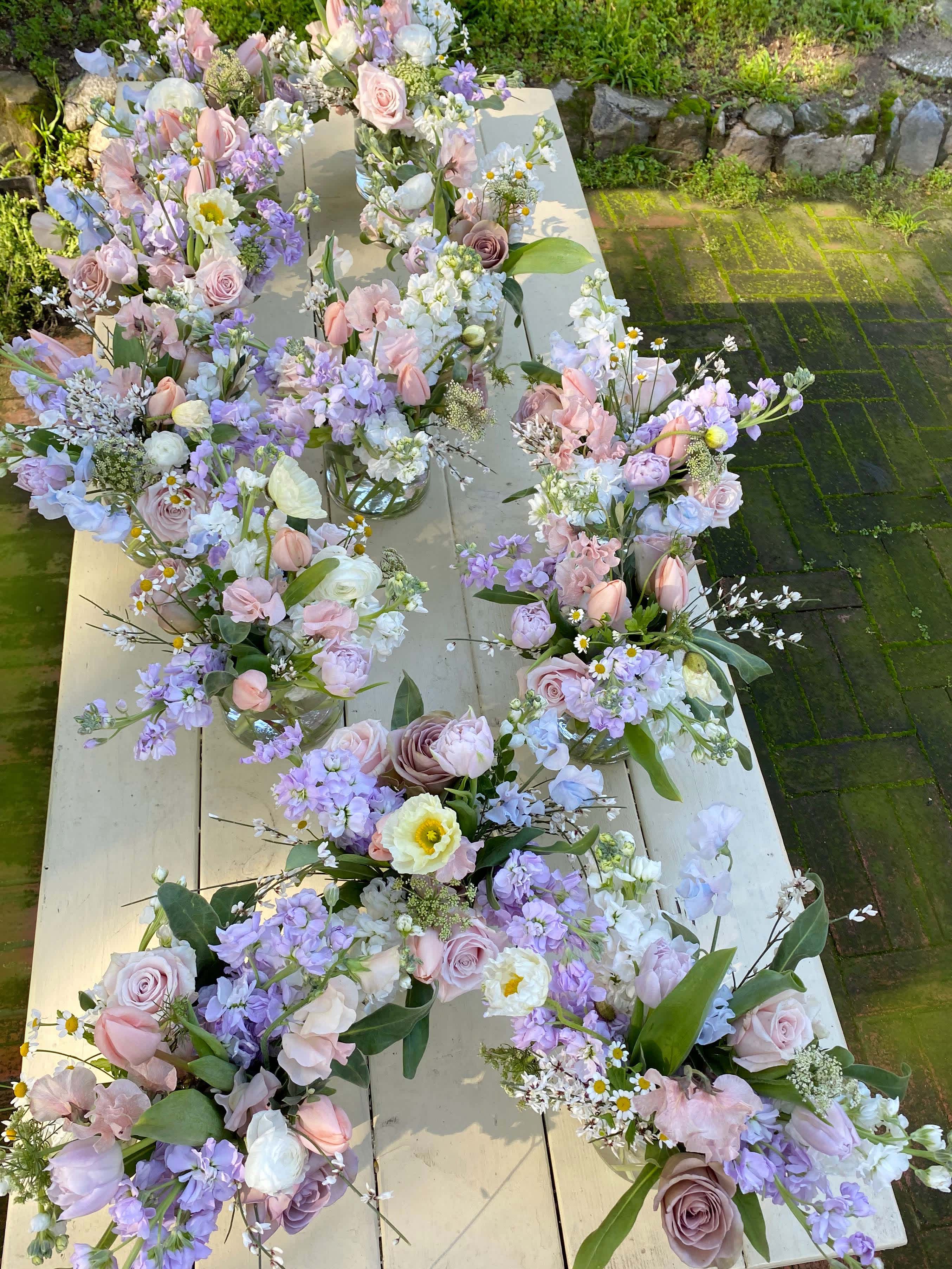 Pastel floral bouquets arranged in small glass vases on a long table outdoors.