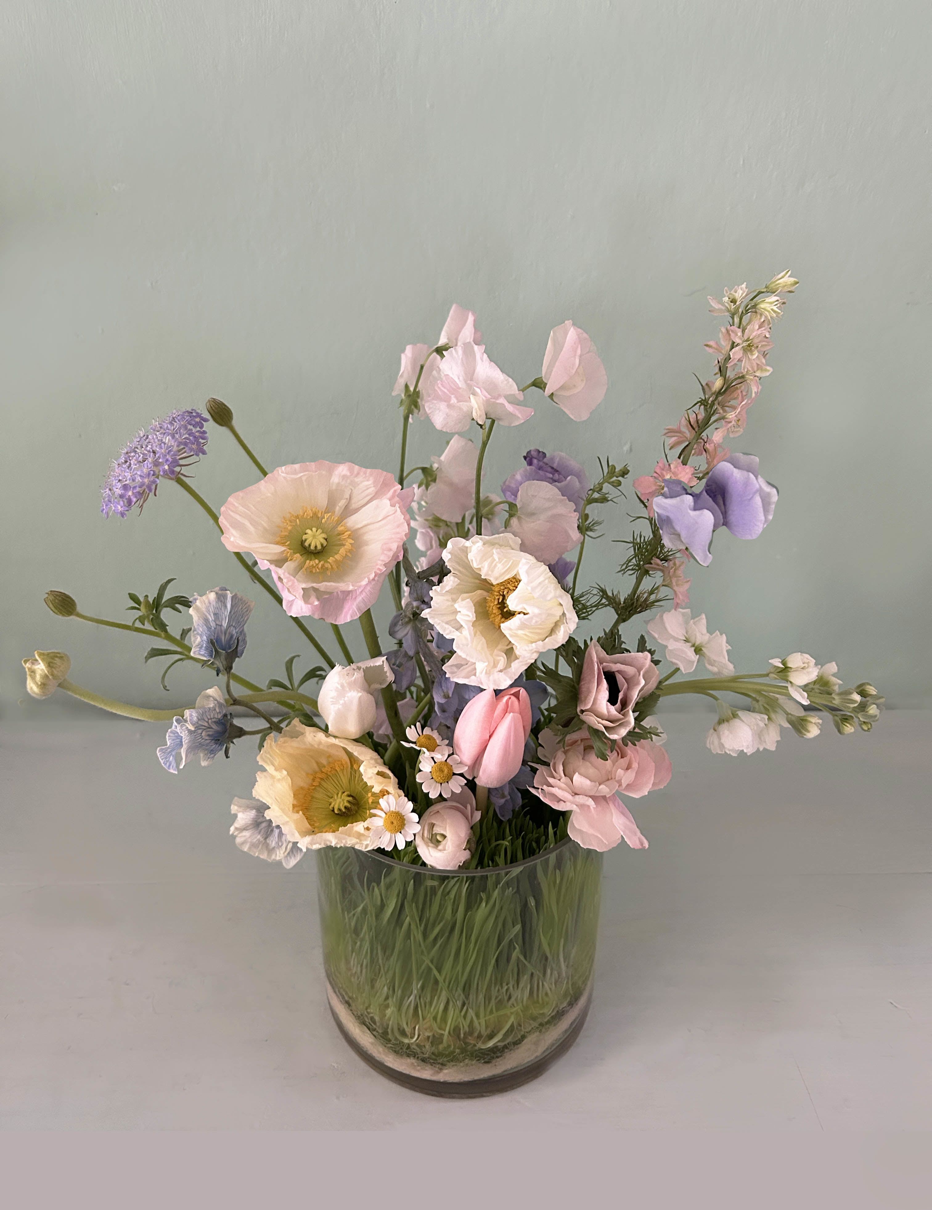 The Love Garden - Bring the garden indoors in this sweet Blomst creation. A petite garden in a vase, with beautiful fresh stems on a nest of grass. Seasonal stems of poppies, ranunculus, sweet peas, tulips etc adorn the clear cylinder vase with grass base. An elegant and timeless floral gift. Add lovely gifts at checkout for a even more thoughtful floral gift.