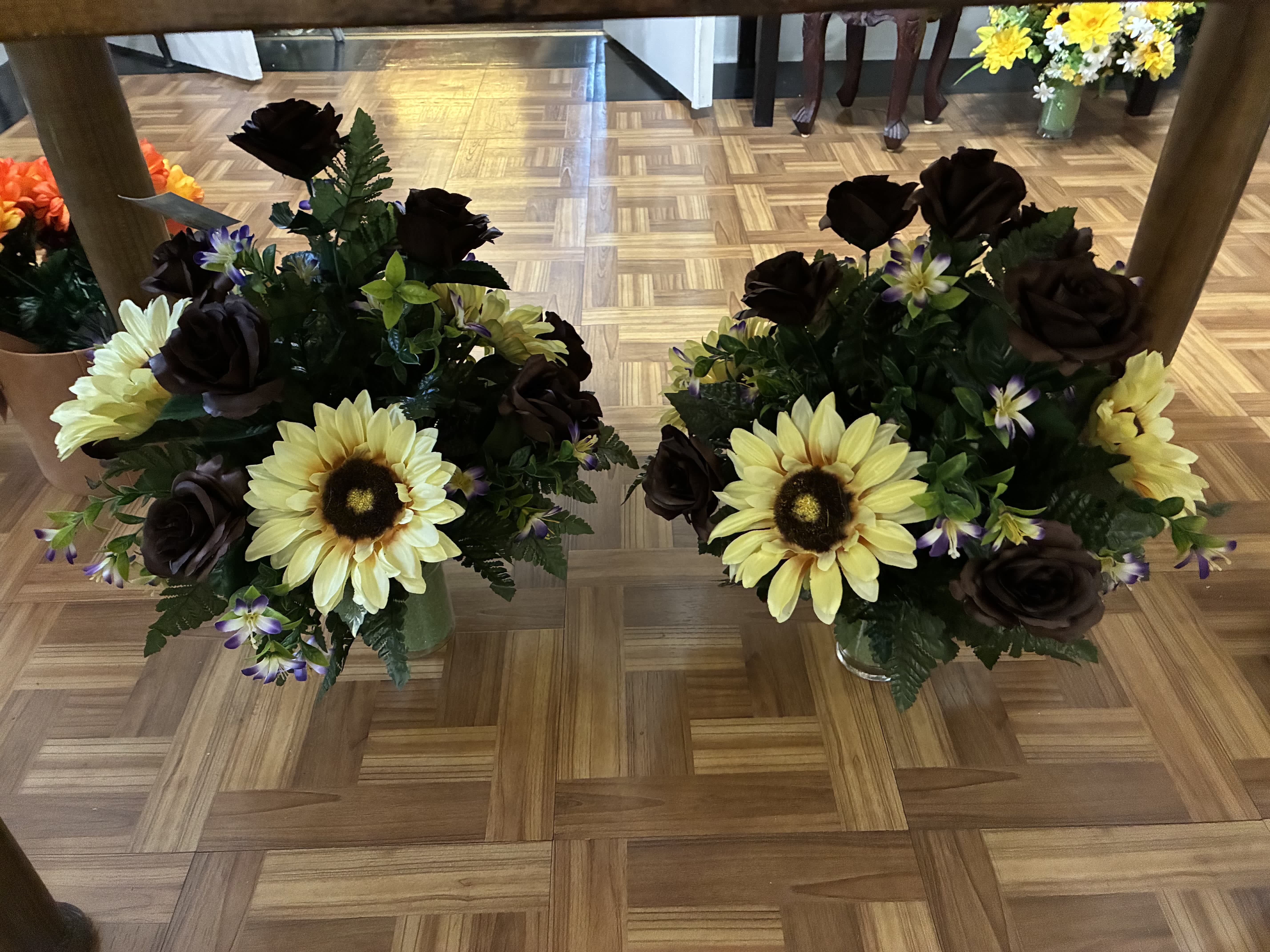 Two floral arrangements with yellow sunflowers and dark roses in glass vases