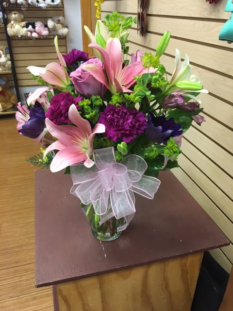 Pink lilies and purple flowers arranged in a glass vase with a sheer ribbon