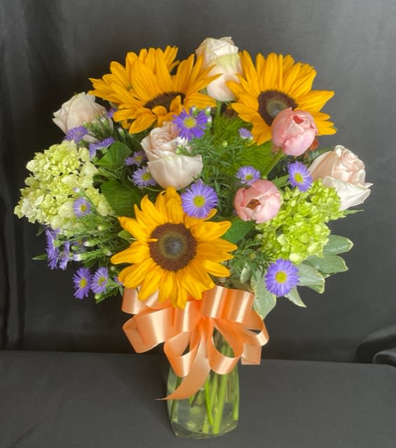 Bouquet of sunflowers, roses, and purple daisies in a glass vase with a peach ribbon