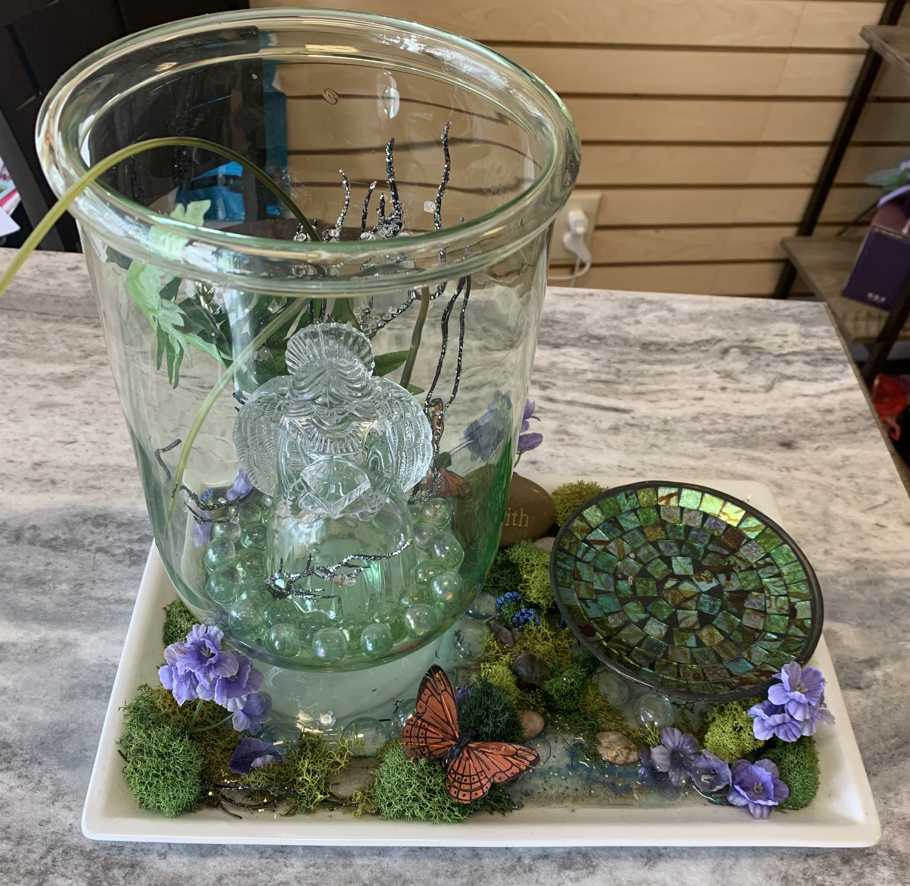 Glass fountain display with purple flowers and butterfly accents on a tray