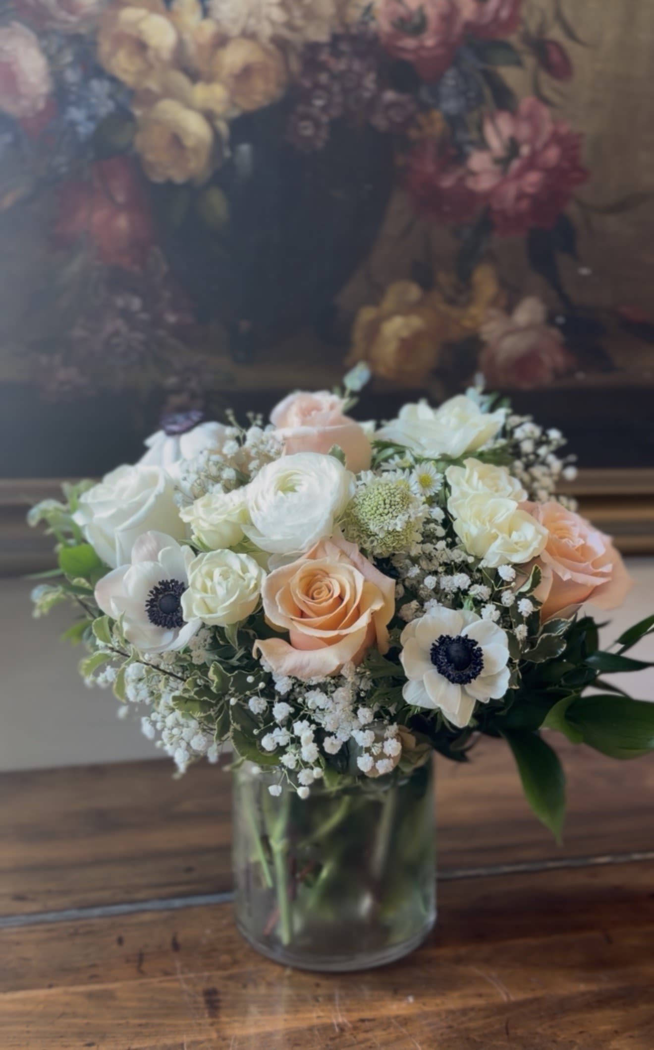 Round arrangement of white and peach roses, ranunculus, and anemones in a clear glass vase.
