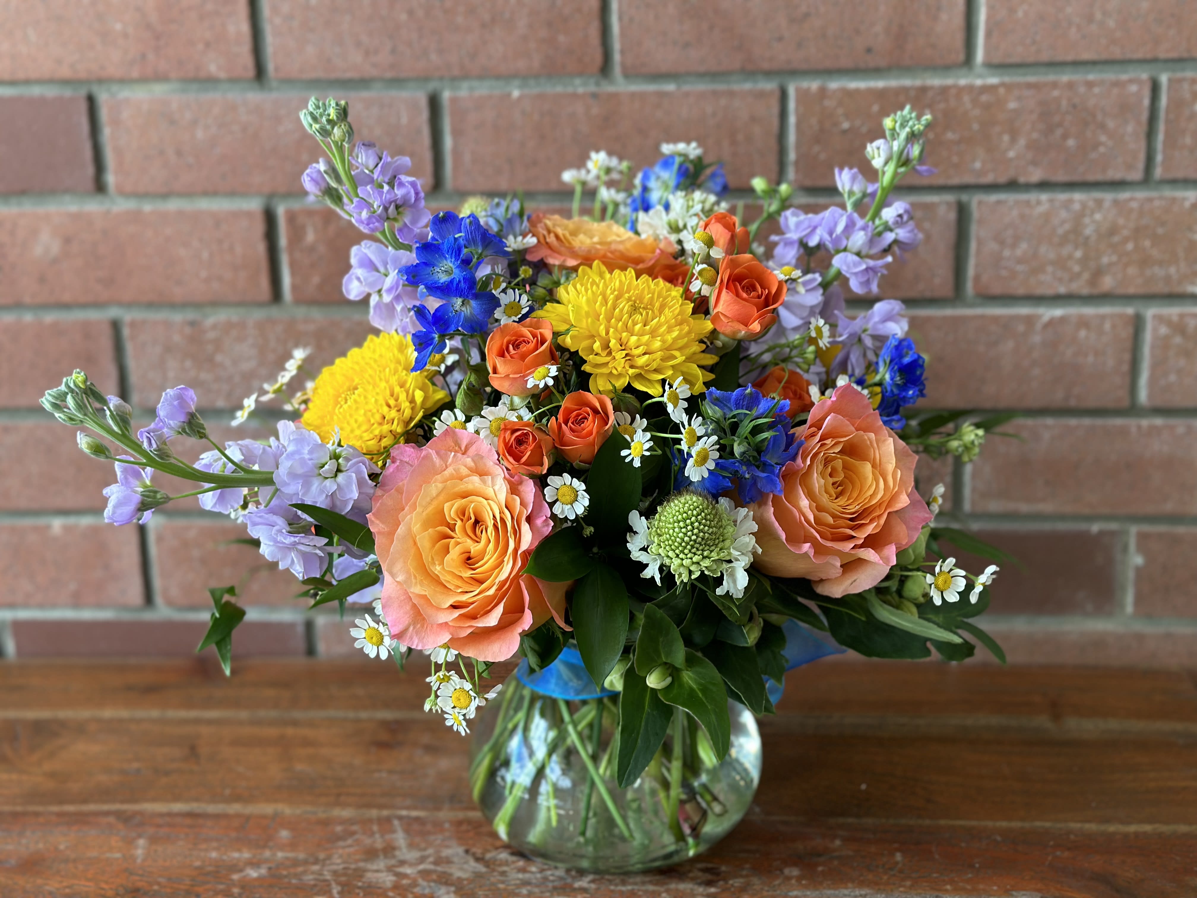Mixed bouquet of orange roses, yellow mums, blue flowers and white daisies in a round glass vase