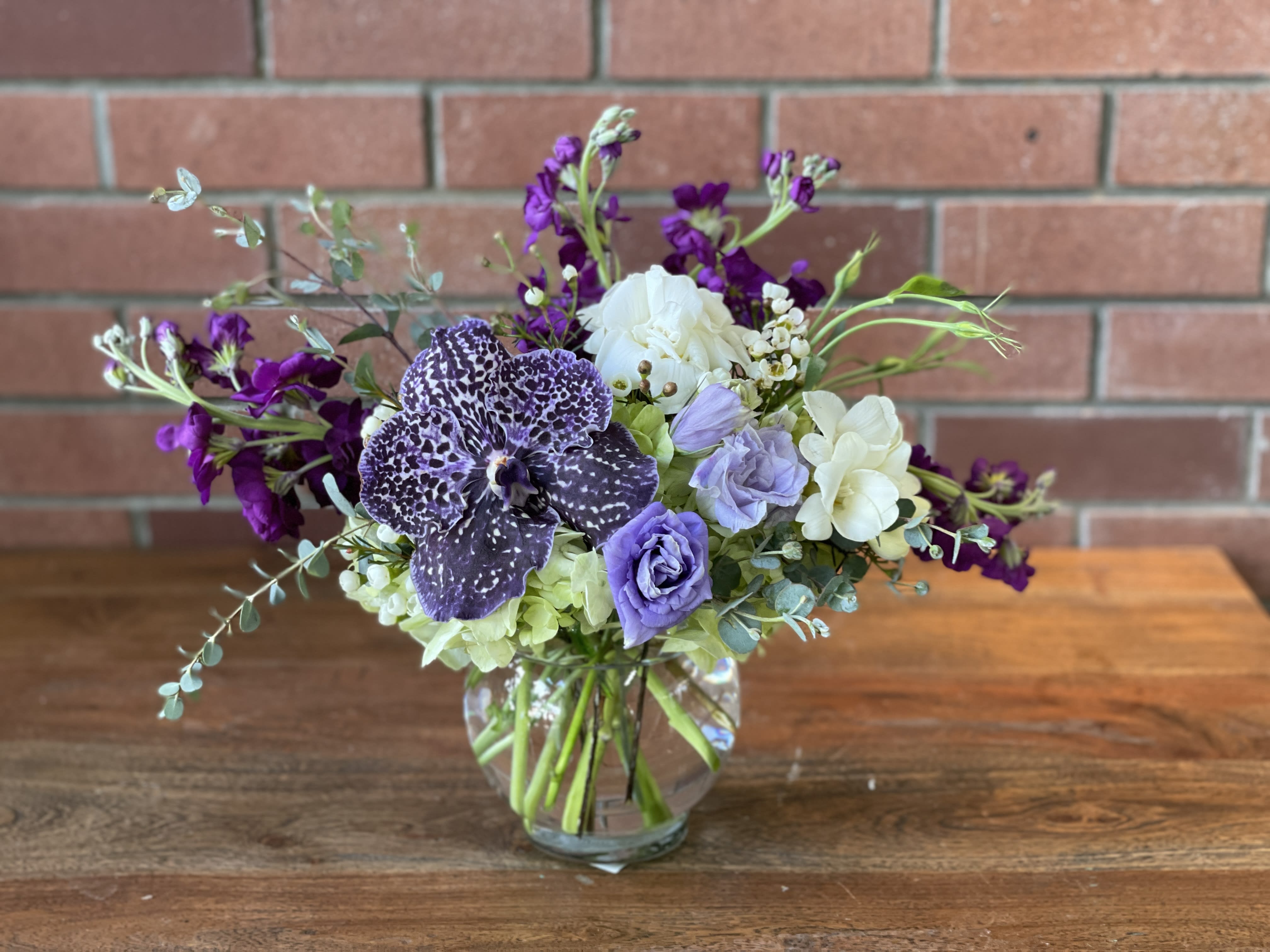Low glass vase with purple orchid, lavender lisianthus, white roses and mixed blooms on a table.