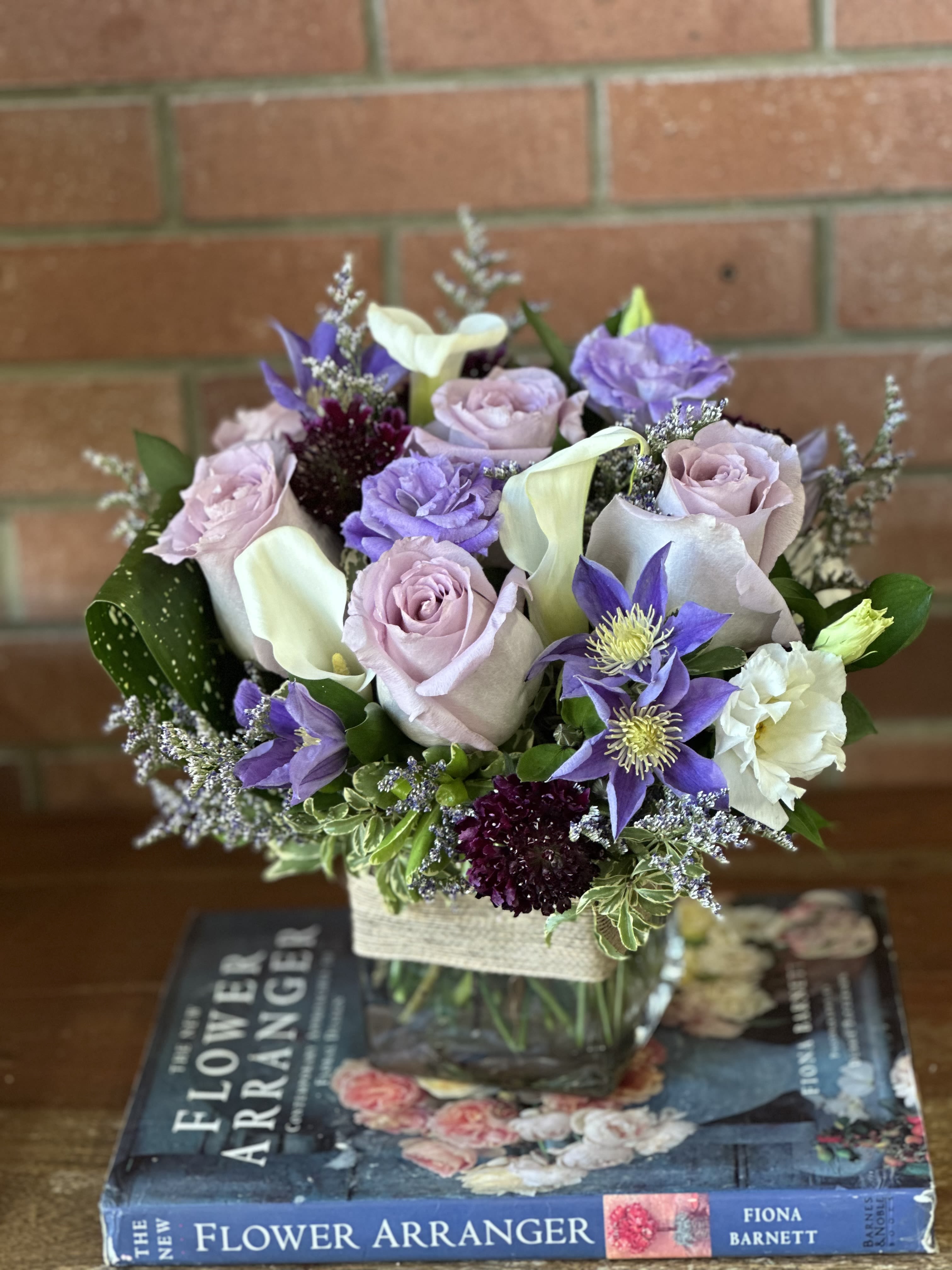Low lavender and white flower arrangement with roses and calla lilies in a glass cube vase on top of books