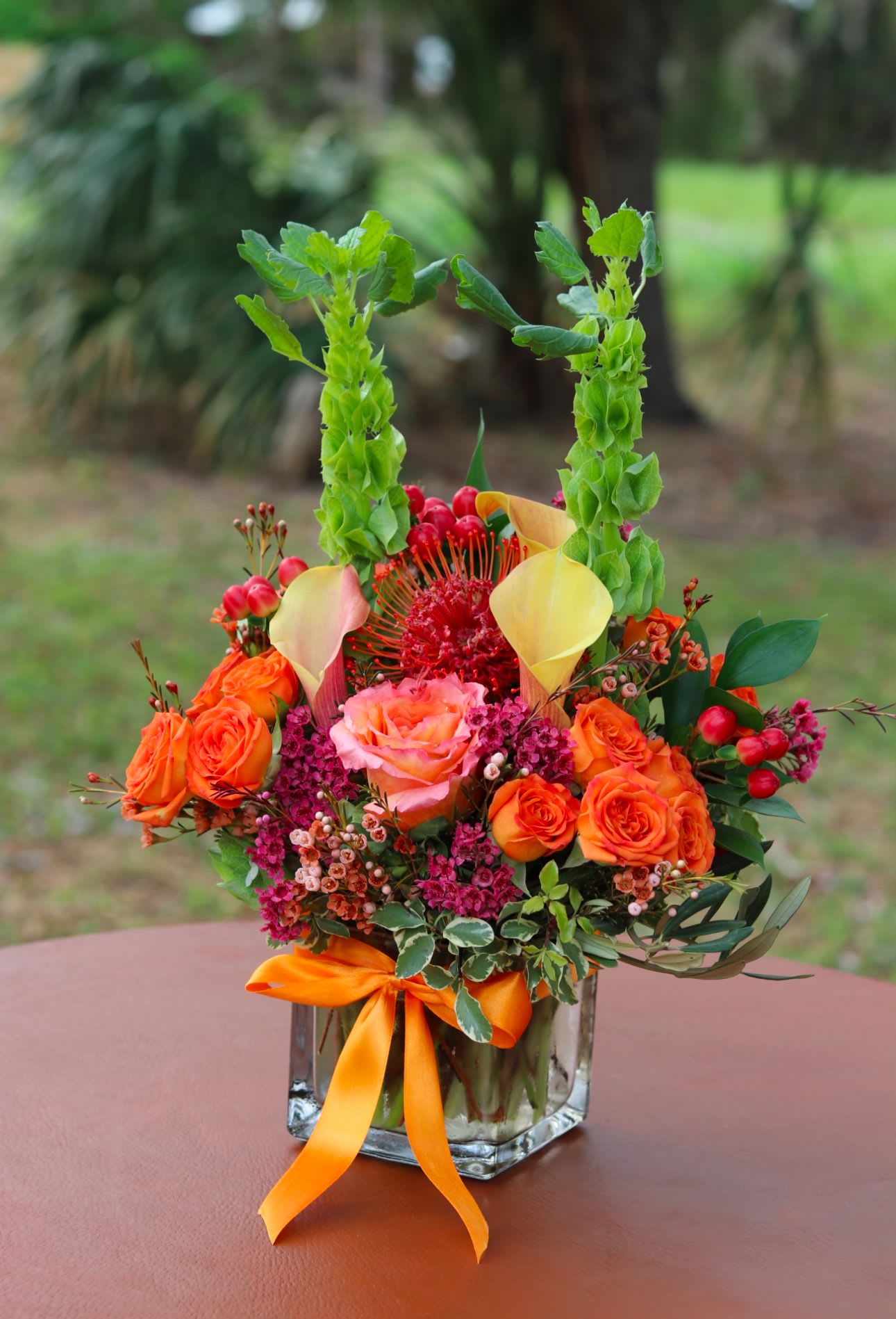 Bright orange and pink rose arrangement with yellow calla lilies and tall green bells of Ireland in a glass vase