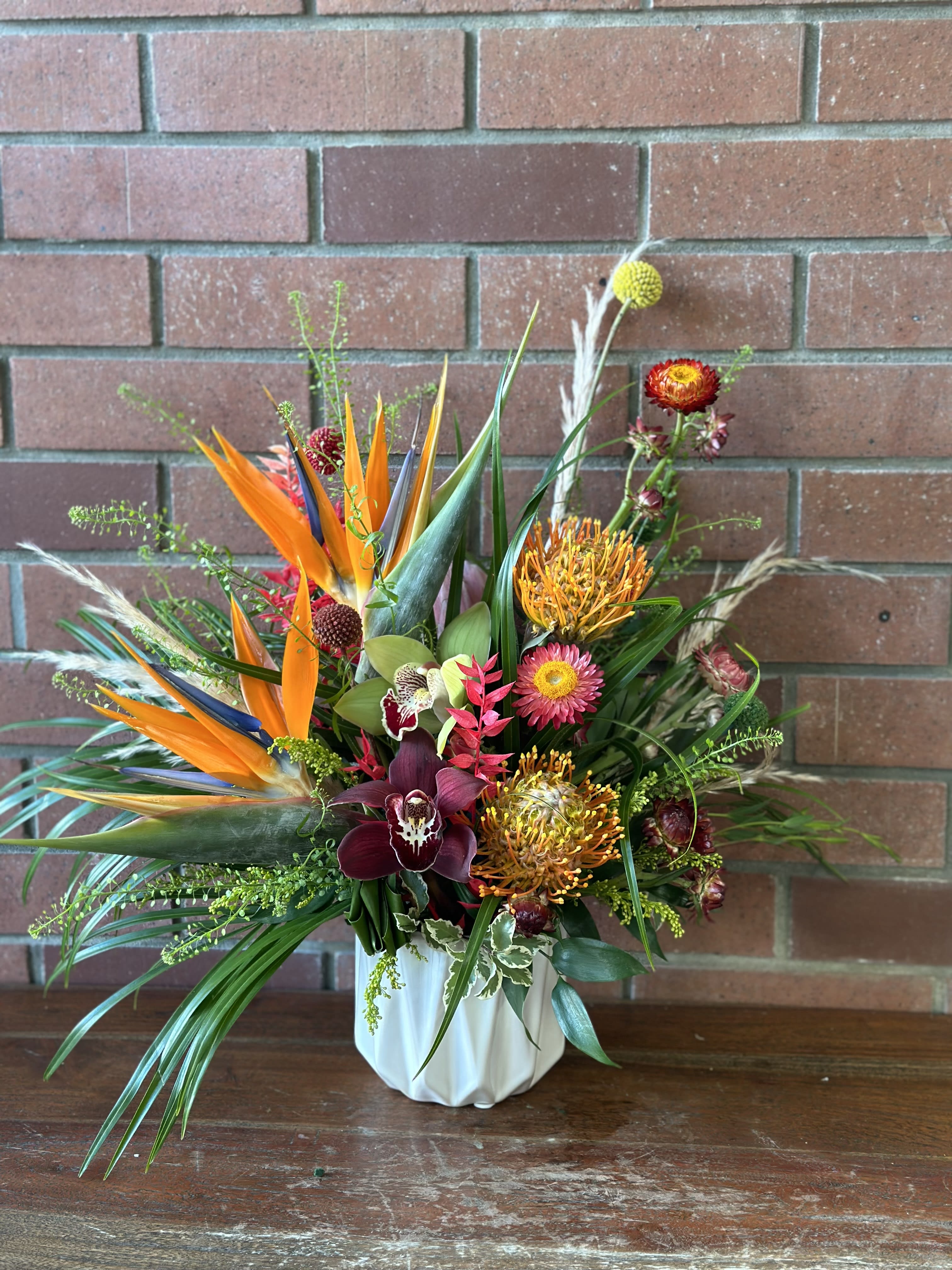 Tropical arrangement with orange birds of paradise, protea, and orchids in a white ceramic vase