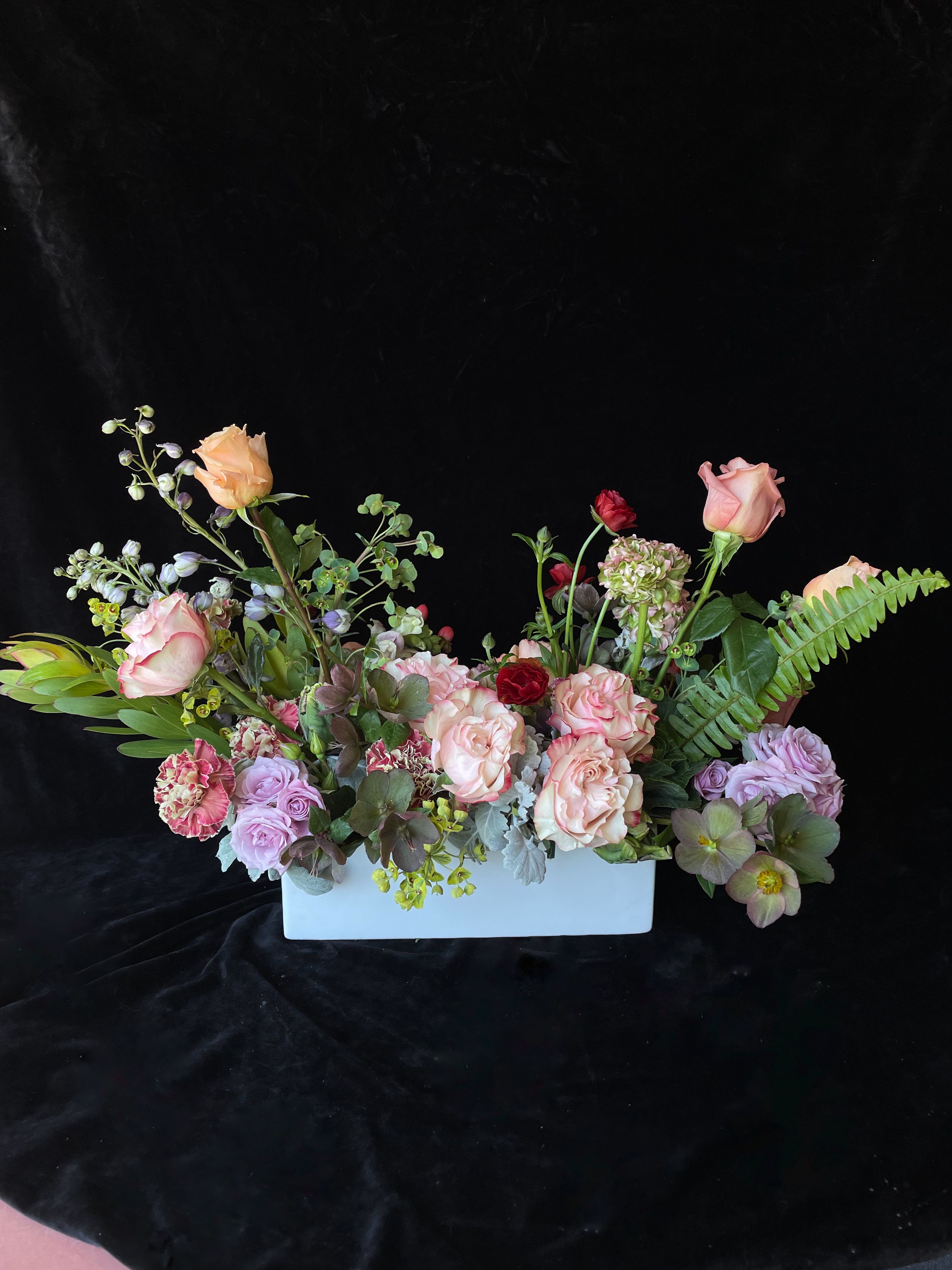 Low pastel arrangement of peach roses, lavender flowers, and green accents in a white rectangular vase