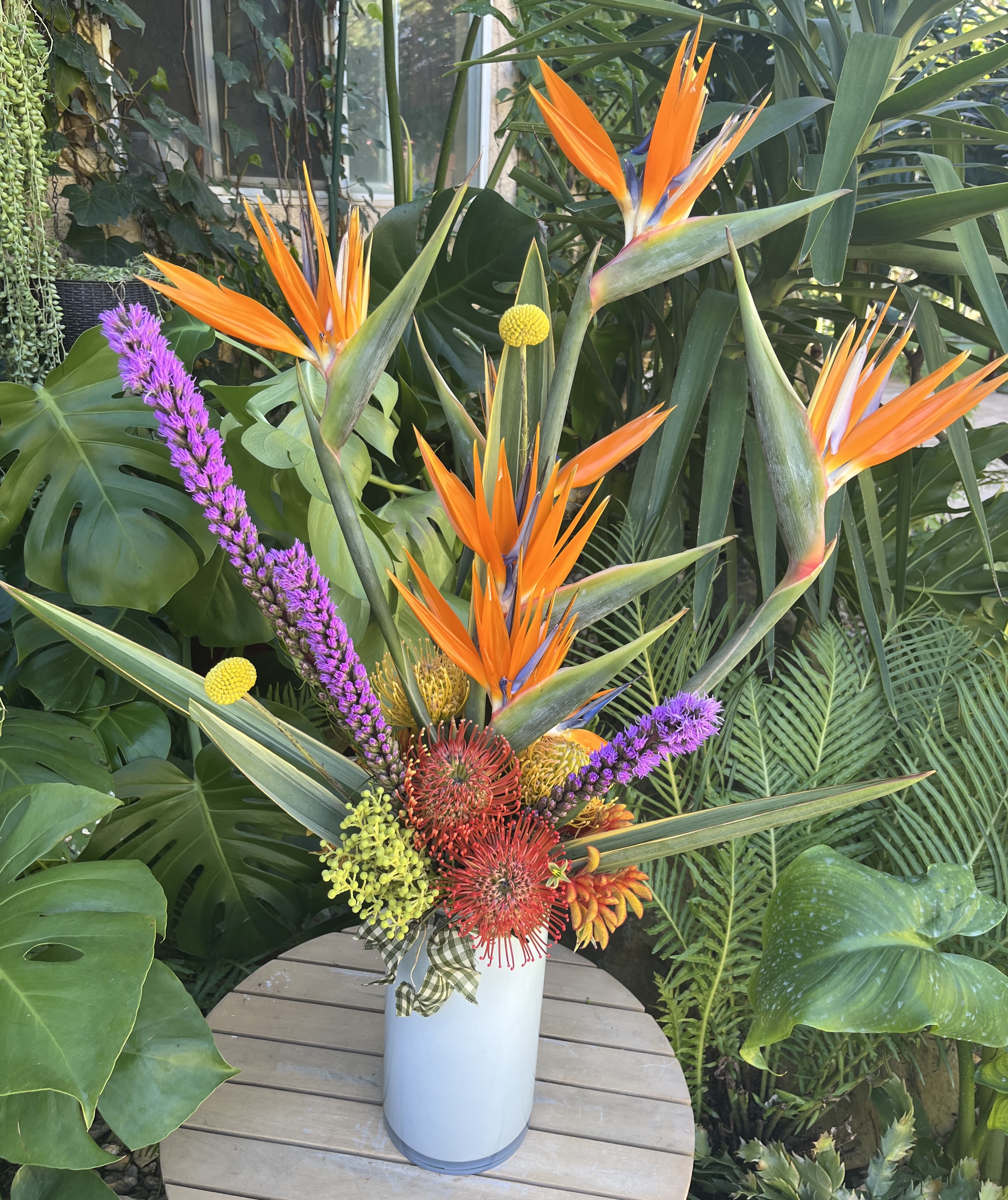 Tropical arrangement with orange bird-of-paradise and purple accents in a white vase