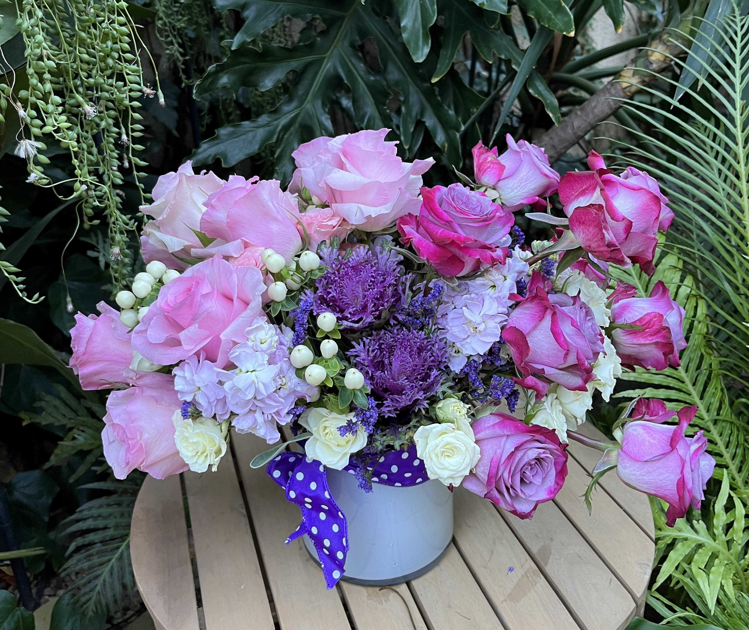 Pink and white roses arranged in a white bucket with a purple ribbon