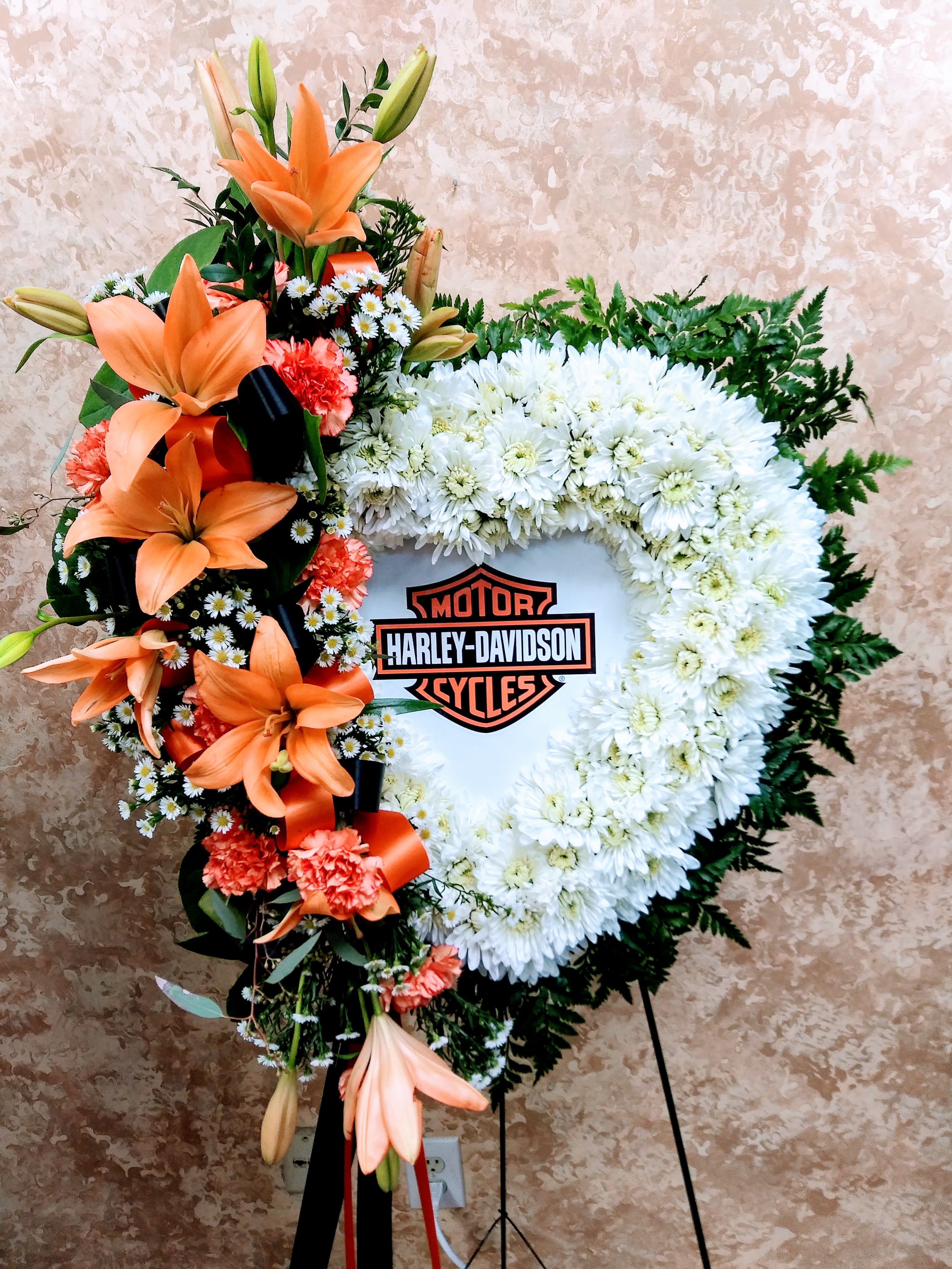 Heart-shaped floral wreath with orange lilies and white chrysanthemums around a Harley-Davidson logo