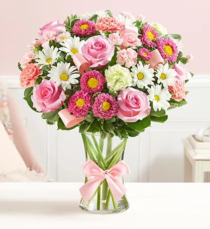 Pink roses and daisies arranged in a clear glass vase with a pink ribbon