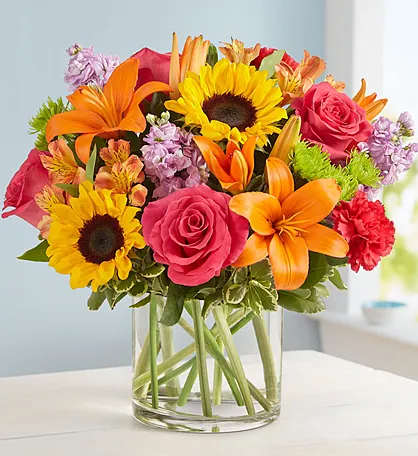 Bright mixed bouquet in a clear glass vase with sunflowers, roses, lilies, and carnations