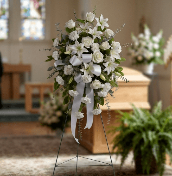 Sentiments of Serenity Spray - This elegant standing spray is a beautiful expression of peace, comfort, and remembrance. Designed with soft yellow lilies, creamy roses, and delicate white carnations, it’s gracefully accented with lush greenery that adds fullness and natural movement. The gentle yellow tones symbolize warmth, hope, and heartfelt memories, while the white blooms reflect purity and serenity. Finished with a classic white ribbon, this spray is a timeless tribute—perfect for honoring a loved one with grace and love.  