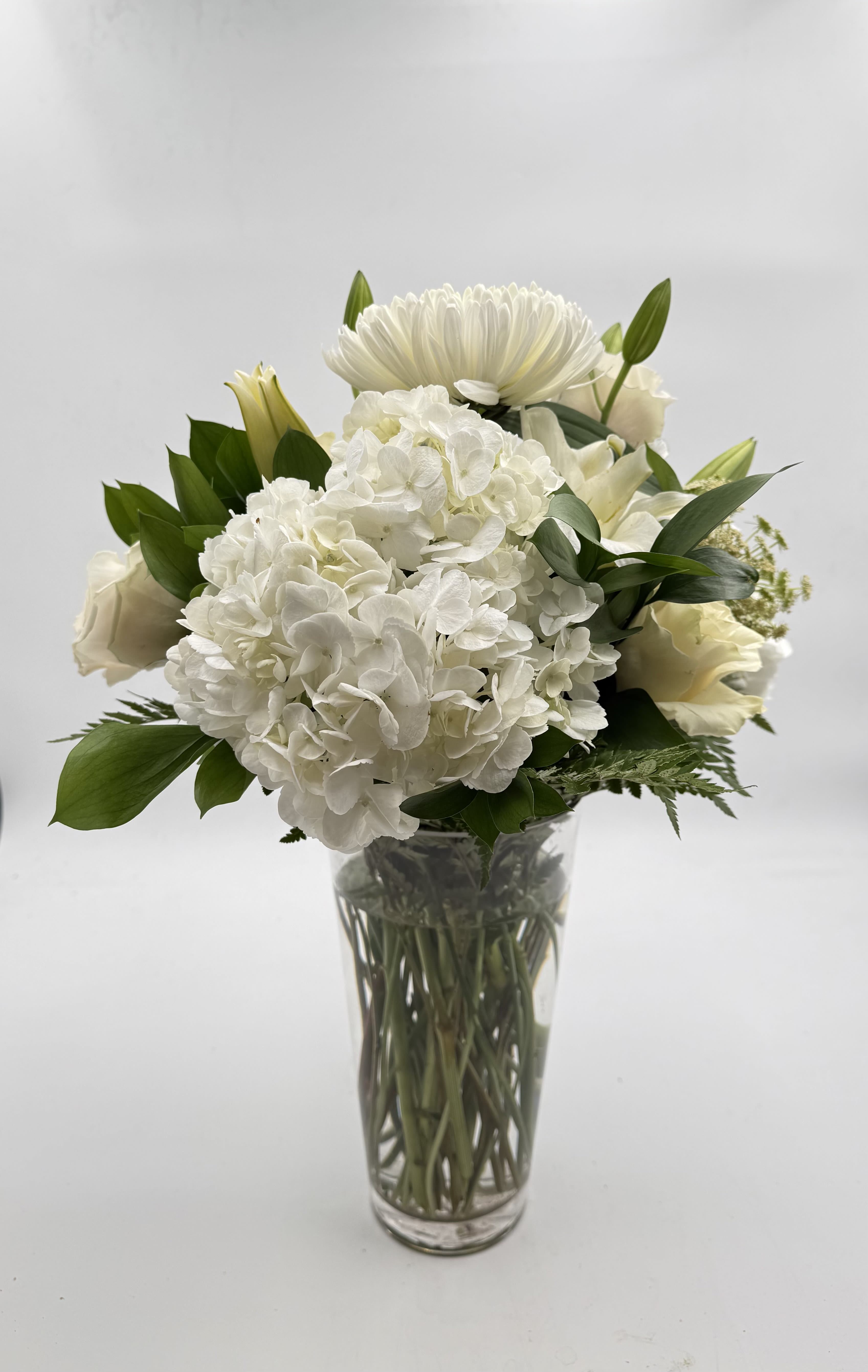 Deepest Sympathy Vase Arrangment - A quiet, composed centerpiece built around a dense cluster of white hydrangeas, this sympathy vase arrangement reads as calm and comforting-the kind of flower arrangement that helps the person you're thinking of feel seen. The rounded profile is topped by a single large white chrysanthemum-like bloom and framed with white lilies and pale cream roses, making it an appropriate choice for sympathy or funeral settings as well as other moments that call for gentle reassurance.  The palette is mostly white with touches of green foliage for contrast; the clear glass vase reveals the stems for an airy, understated look that complements traditional services or a simple bedside sympathy gift. Textures move from the soft mophead hydrangeas to the sculpted rose petals and the vertical presence of the chrysanthemum, creating a layered, elegant composition without being fussy.  Choose this arrangement when you want to convey reverence and calm-for memorial services, to comfort someone at home, or as a refined, white wedding-adjacent centerpiece. The compact sizing fits easily on tabletops or display stands; to keep blooms fresh, replace vase water every two days and trim stems slightly when you refresh the water.