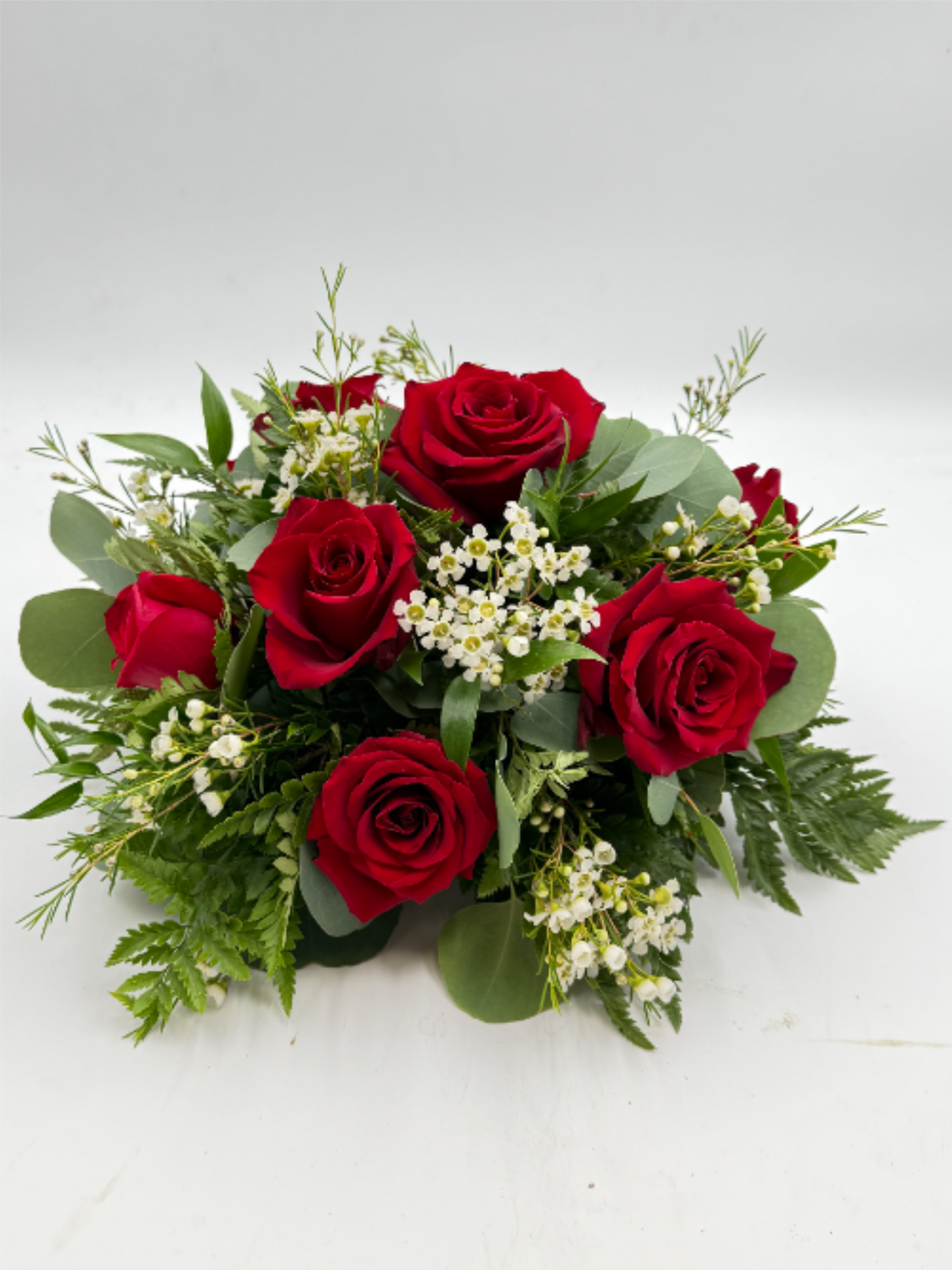 Classic Rose Centerpiece - Deep clusters of deep red roses form the heart of this low, rounded flower arrangement-perfect as a romantic anniversary roses centerpiece or a formal wedding table arrangement. The rich red-and-white palette reads classic and elegant, so the person you're thinking of will feel truly celebrated whether this is set at a dinner table, used as a sympathy tribute, or placed as a holiday focal point.  Small white filler blossoms are woven between the roses and framed by a mix of glossy and matte greens and fern-like foliage, giving the design a full, layered texture without rising tall above the table. The compact, medium hand-tied shape keeps blooms close to the surface so guests can converse across it; it looks polished on a white tablecloth alongside wine glasses, dinner plates, and neatly folded napkins.  Ideal for weddings, anniversaries, formal dinners, or a quiet yet meaningful gift, this centerpiece sends a message of timeless affection and refined taste. The balanced, neat presentation needs only basic care: keep stems in fresh water, trim occasionally, and swap water every few days to maintain that lush, just-arranged look through the celebration.