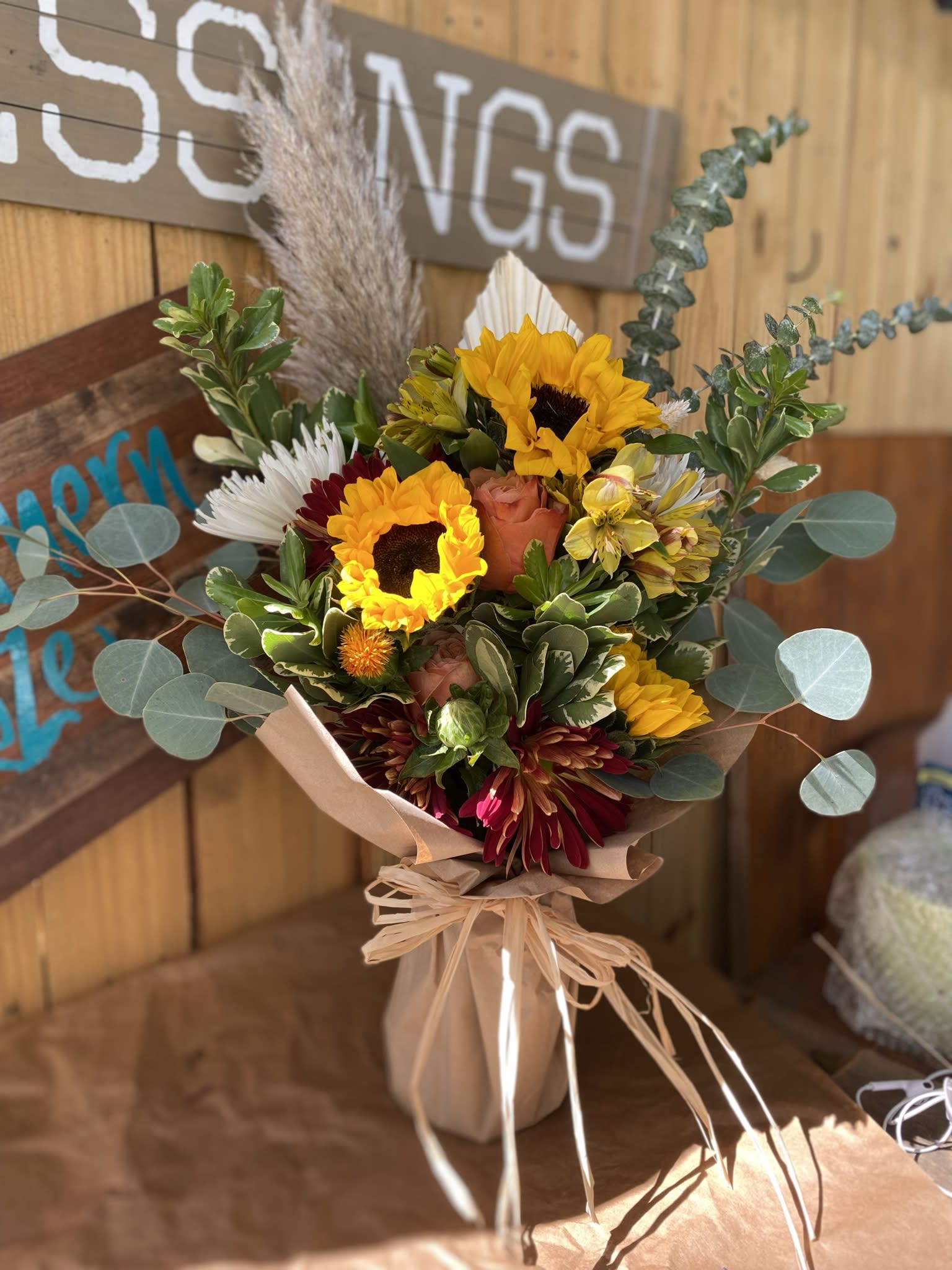 Rustic bouquet of yellow sunflowers, orange roses, and burgundy blooms wrapped in brown paper and raffia.