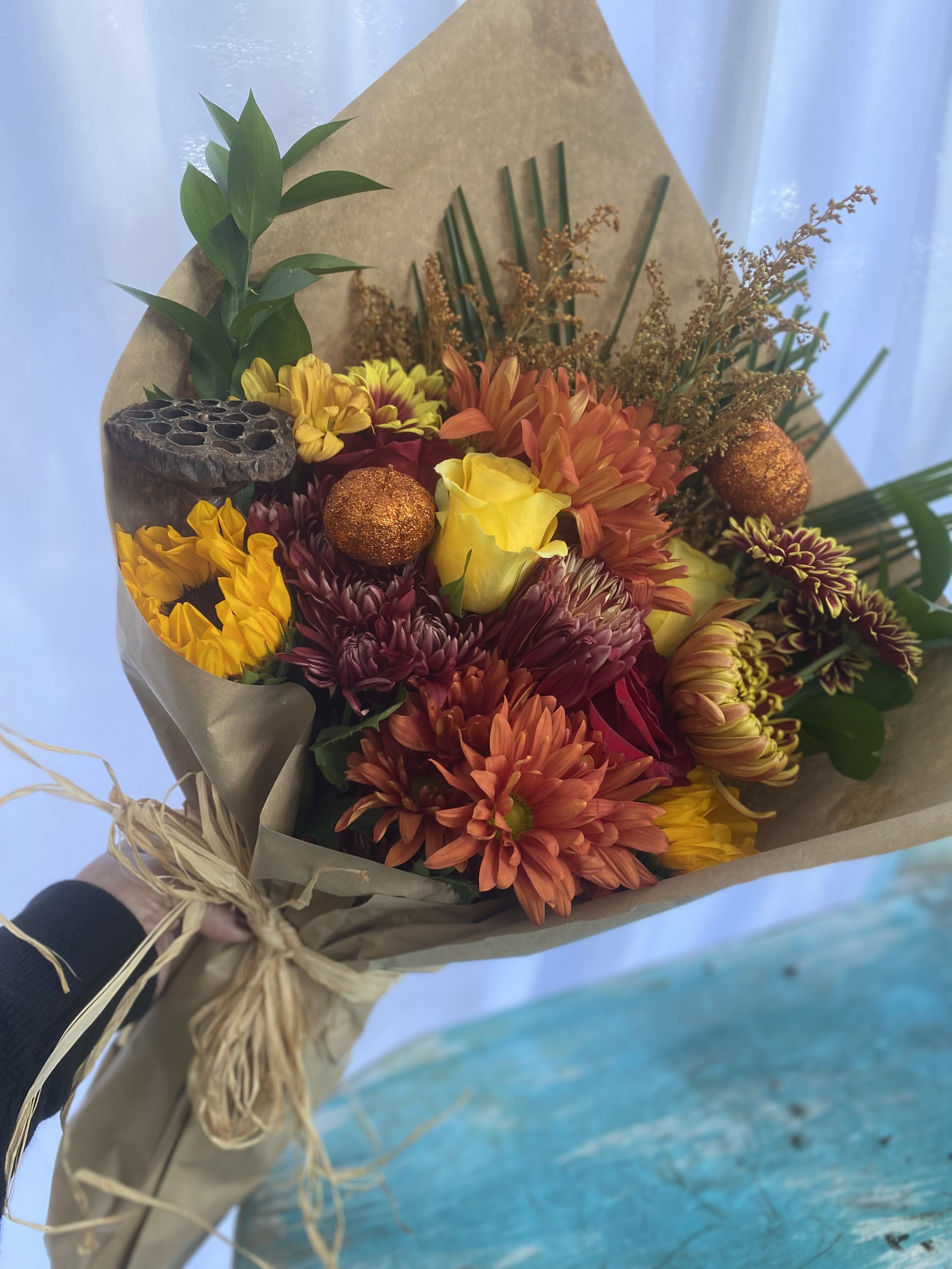 Hand-tied bouquet of orange mums and yellow roses wrapped in kraft paper with a raffia bow