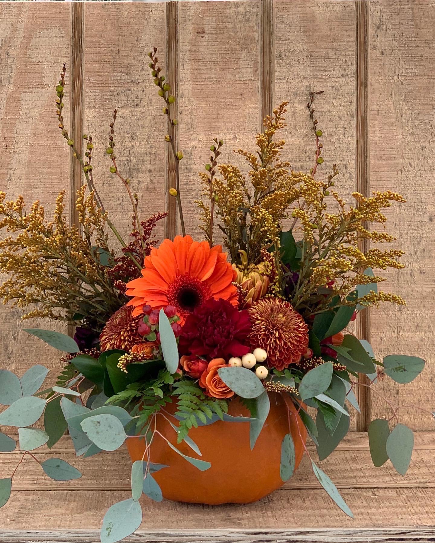 Autumn arrangement of orange gerbera, roses, and mums in a pumpkin container with eucalyptus accents