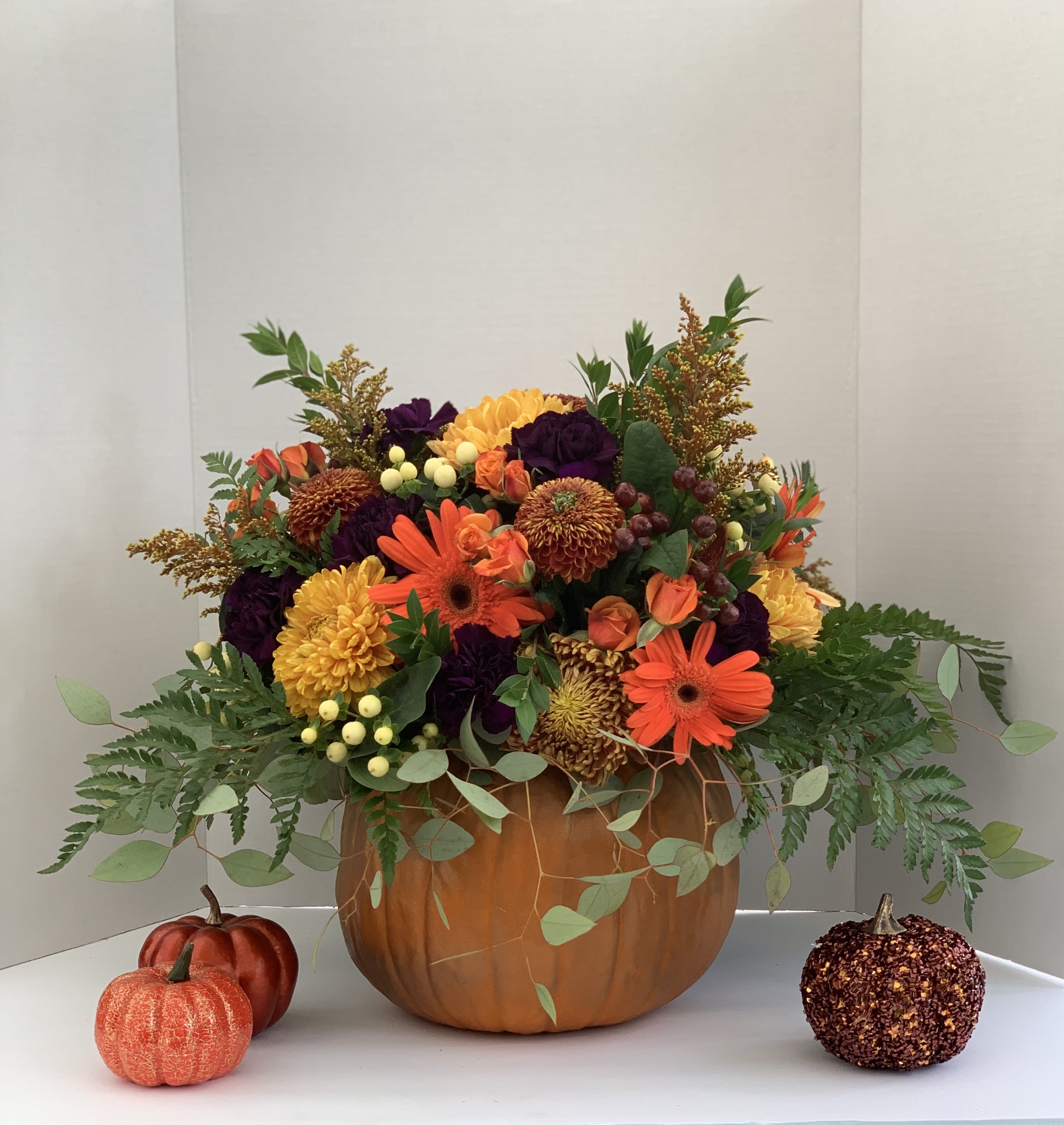 Autumn bouquet of orange and yellow flowers in a pumpkin container with small decorative pumpkins nearby.