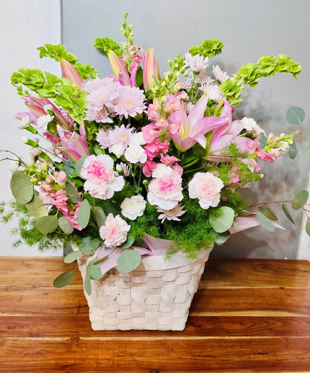 Cotton Candy Dreams - Pink lilies and soft pink-and-white carnations create a gentle, cheerful statement in this spring bouquet - a perfect birthday bouquet or Mother's Day gift that feels fresh and feminine. Tall green snapdragon spikes lift the arrangement, giving it graceful height and a garden-style silhouette that makes the recipient feel truly celebrated and seen.  Arranged in a white woven basket and finished with a pink ribbon, the mix includes airy eucalyptus-like foliage that adds texture and a lively, layered look. The pastel pinks, clean white accents, and verdant greens work together for a calming yet joyful impression; the basket measures as a compact tabletop size ideal for dining tables, desks, or kitchen counters. This is a thoughtful way to mark birthdays, Mother's Day, or a springtime surprise - simple care: keep in fresh water and out of direct heat to extend blooms.