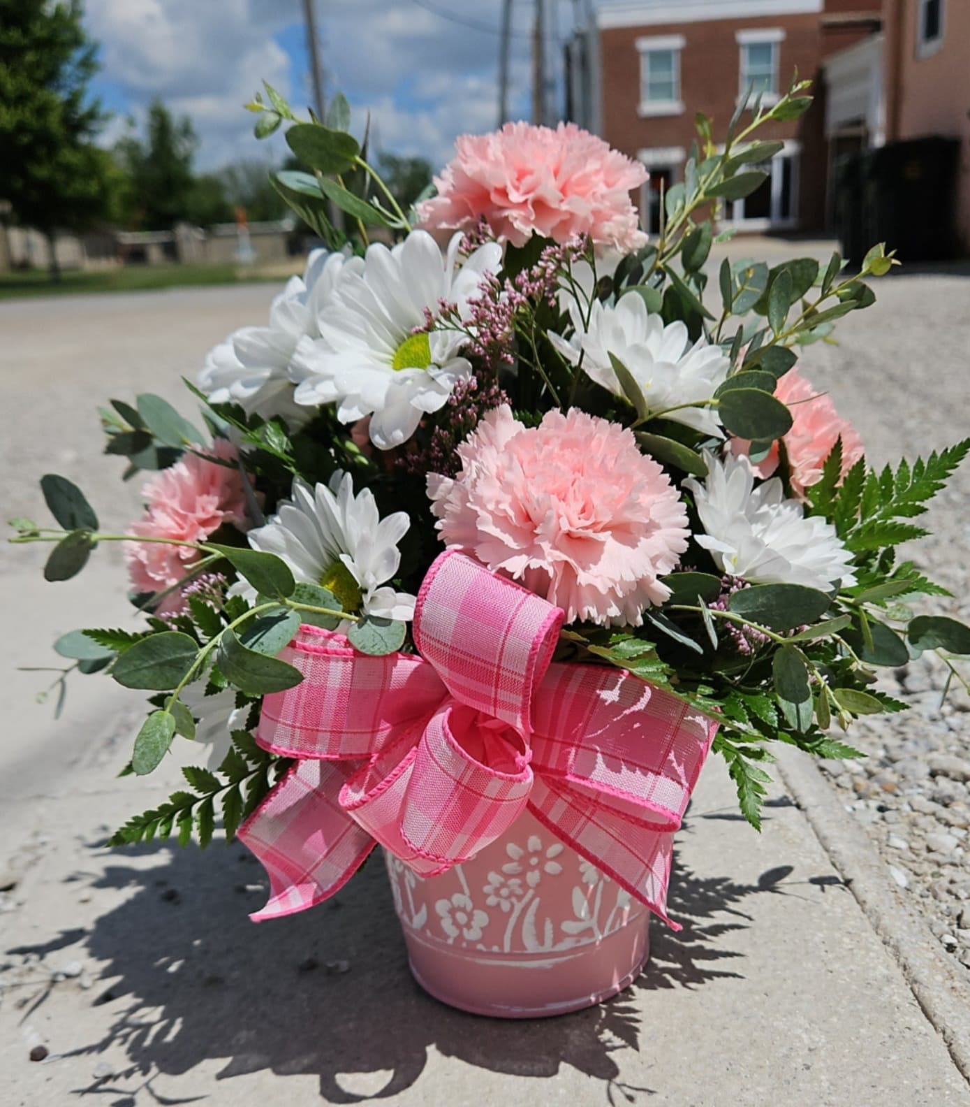 Daisies &amp; Carnations  - Soft pink carnations paired with crisp white daisies create a fresh, garden-fresh bouquet that feels cheerful and kindly - perfect as a birthday bouquet, a thank-you arrangement, or a simple everyday gift to brighten someone's day. The compact, rounded design presents a soft, feminine look that reads as both casual and thoughtfully crafted for the person you're thinking of.  The arrangement sits in a sweet pink tin bucket and is finished with a large pink plaid ribbon for a handcrafted, gift-ready presentation. Airy greenery and small filler sprigs tuck between the blooms to add texture and a light, natural movement; colors are pink, white, and lively green for a springlike palette. Sized to be a charming centerpiece or bedside surprise, this is the kind of bouquet that makes someone feel seen and celebrated without fuss - easy to order and ready to gift.
