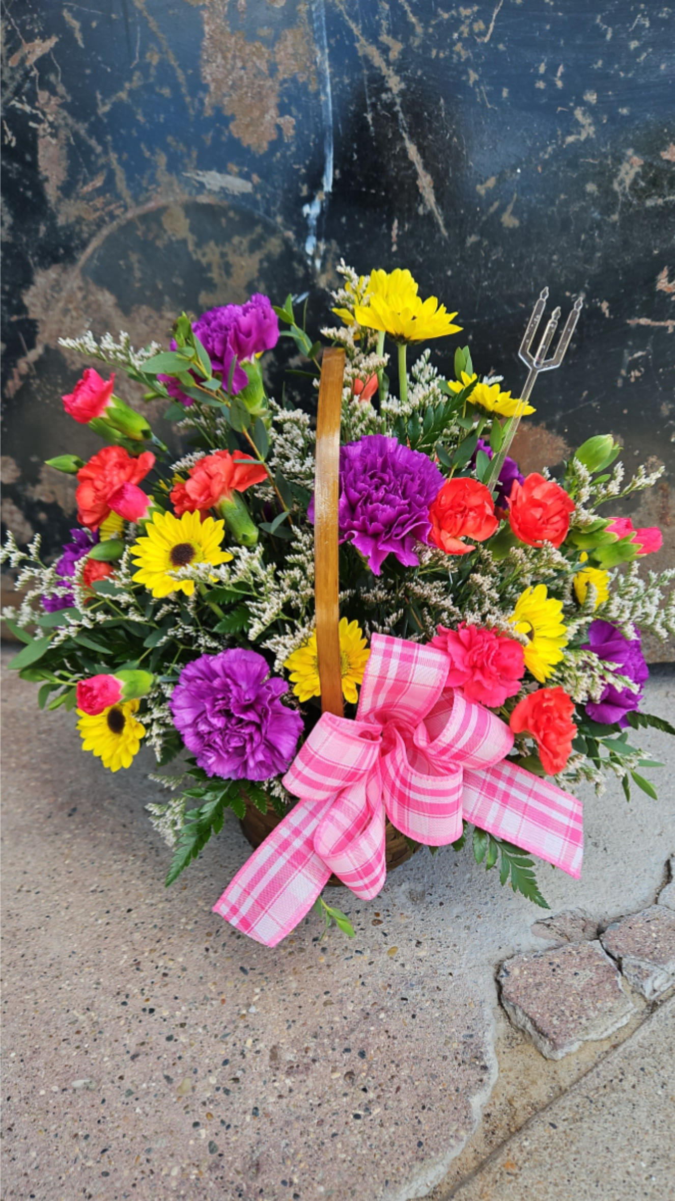 A Basket Full of Colors - A woven basket overflowing with vivid pink, purple, red and sunny yellow blooms-this bright flower arrangement brings instant cheer as a birthday bouquet, congratulations gift, or just-because surprise. The rounded, abundant shape reads lively and informal, so the person you're thinking of will feel celebrated and uplifted the moment they see it.  Packed low in a rustic basket with a wooden handle, the design features vivid carnations and yellow daisy-like blooms layered with airy white filler and mixed greenery for texture and movement. A large pink plaid ribbon tied at the front gives a playful, gift-like finish while plastic floral picks and a plastic trident pick keep stems secure for delivery. The palette and dense, joyful composition make it perfect for sending smiles across many occasions.  Ideal for brightening a kitchen table, office desk, or bedside, this arrangement is an easy, heartfelt way to say happy birthday, congratulations, or simply "I'm thinking of you." No special care is required beyond fresh water-place in a cool spot away from direct sunlight to extend bloom life-and the basket presentation arrives ready to display.
