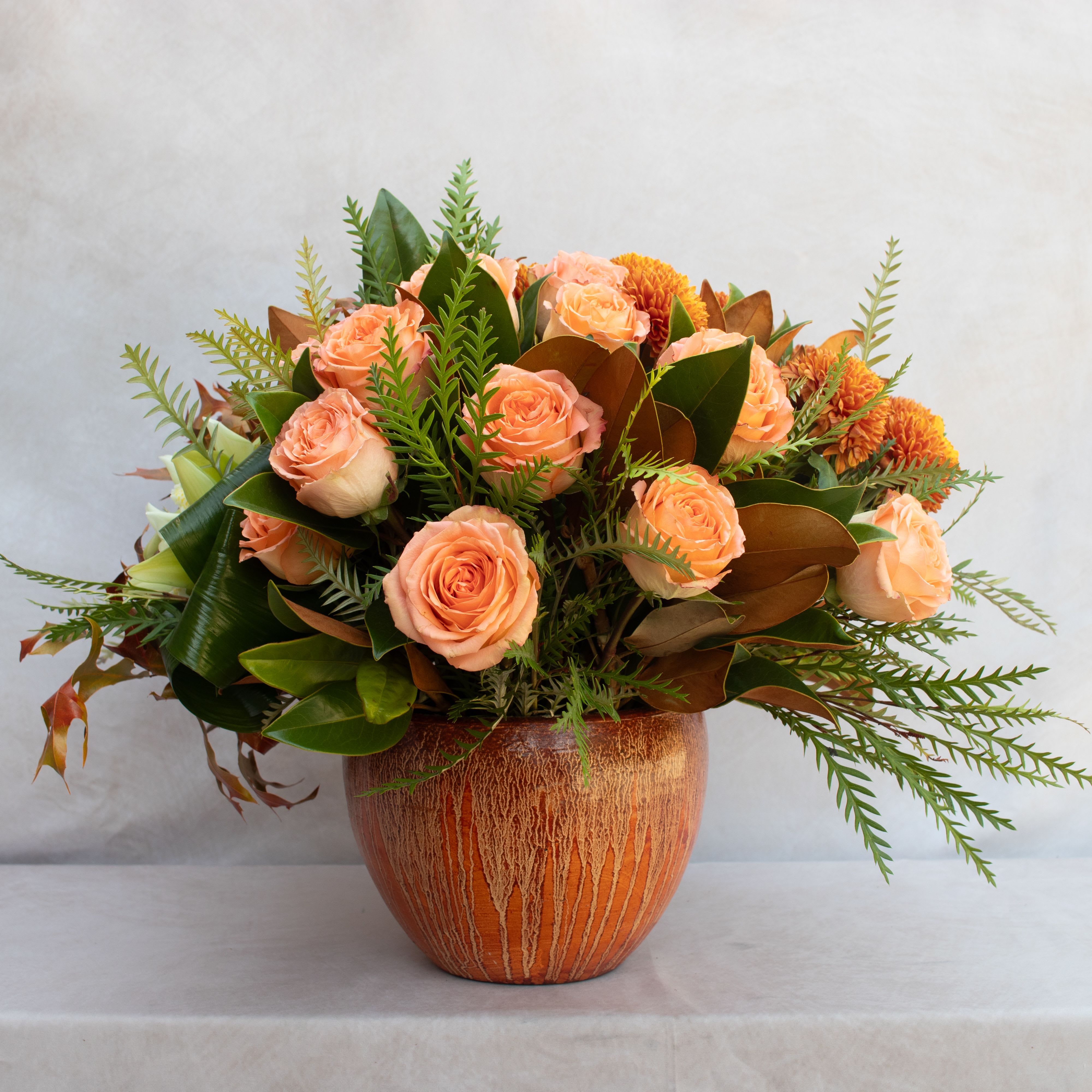TERRACOTTA POTTA  - PEACH ROSES, FALL MUMS, &amp; KALE WITH UNIQUE GREENERY LIKE MAGNOLIA, GREVILLIA, &amp; EUCALYPTUS IN A GLAZED TERRACOTTA POT.  ARRANGEMENT DESIGNED IS ABOUT 18X18. 