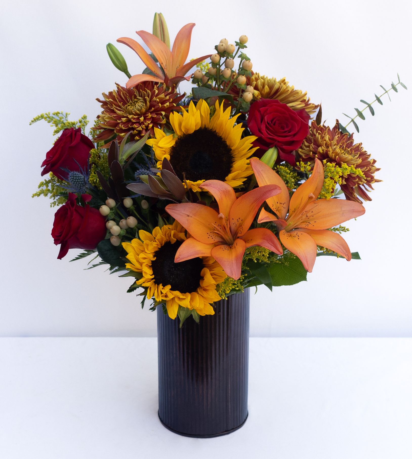 Grand Vibrant Fall Vase   - A large, luxurious bouquet arranged with a lovely mix of fall-colored lilies, protea, sunflowers, cremons, roses, mums, berries, greenery, and fillers designed in a tall, large metal corrugated vase. The Standard vase is 7hx4. The designed arrangement is about 20hx12w. 