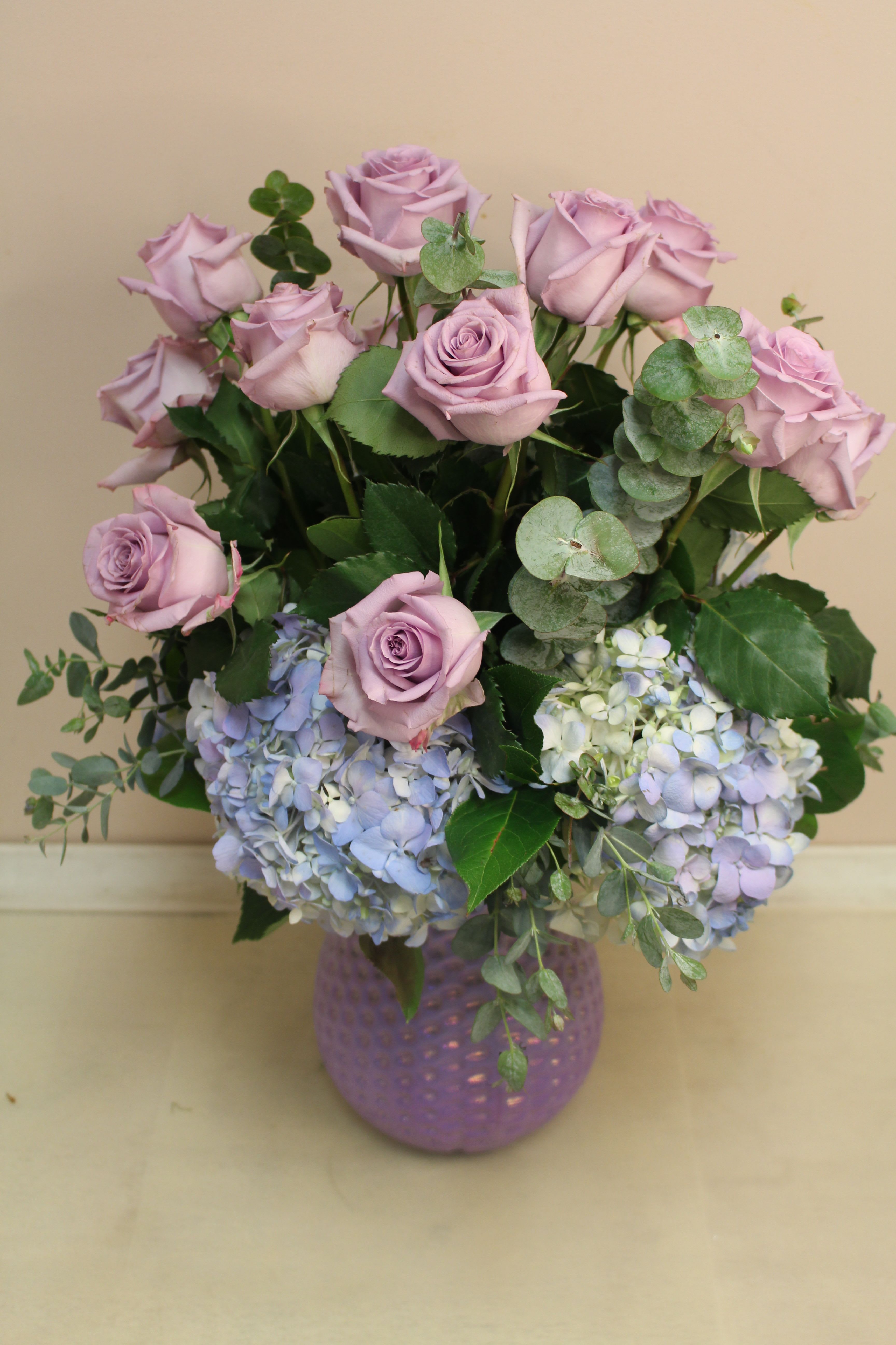 Roses and hydrangea lavender - Soft lavender roses rising above a cloud of pale blue and white hydrangeas create a calm, romantic mood - perfect as a birthday bouquet, anniversary flowers, or a Mother's Day gift. This flower arrangement pairs those lavender roses and lush hydrangeas in a feminine, modern composition that also suits graduations, get well wishes, housewarmings, sympathy, or simply saying "I'm thinking of you."  A round, textured purple glass vase echoes the blooms and gives the design a cohesive, elegant finish. Rounded eucalyptus stems add cool green texture and gentle contrast to the lilac, pale blue and white palette. The arrangement is layered and full, with taller roses forming a refined dome above the hydrangea base for a balanced, refined silhouette - an ideal choice for romance, congratulations, thank-you gestures, or thoughtful everyday moments.