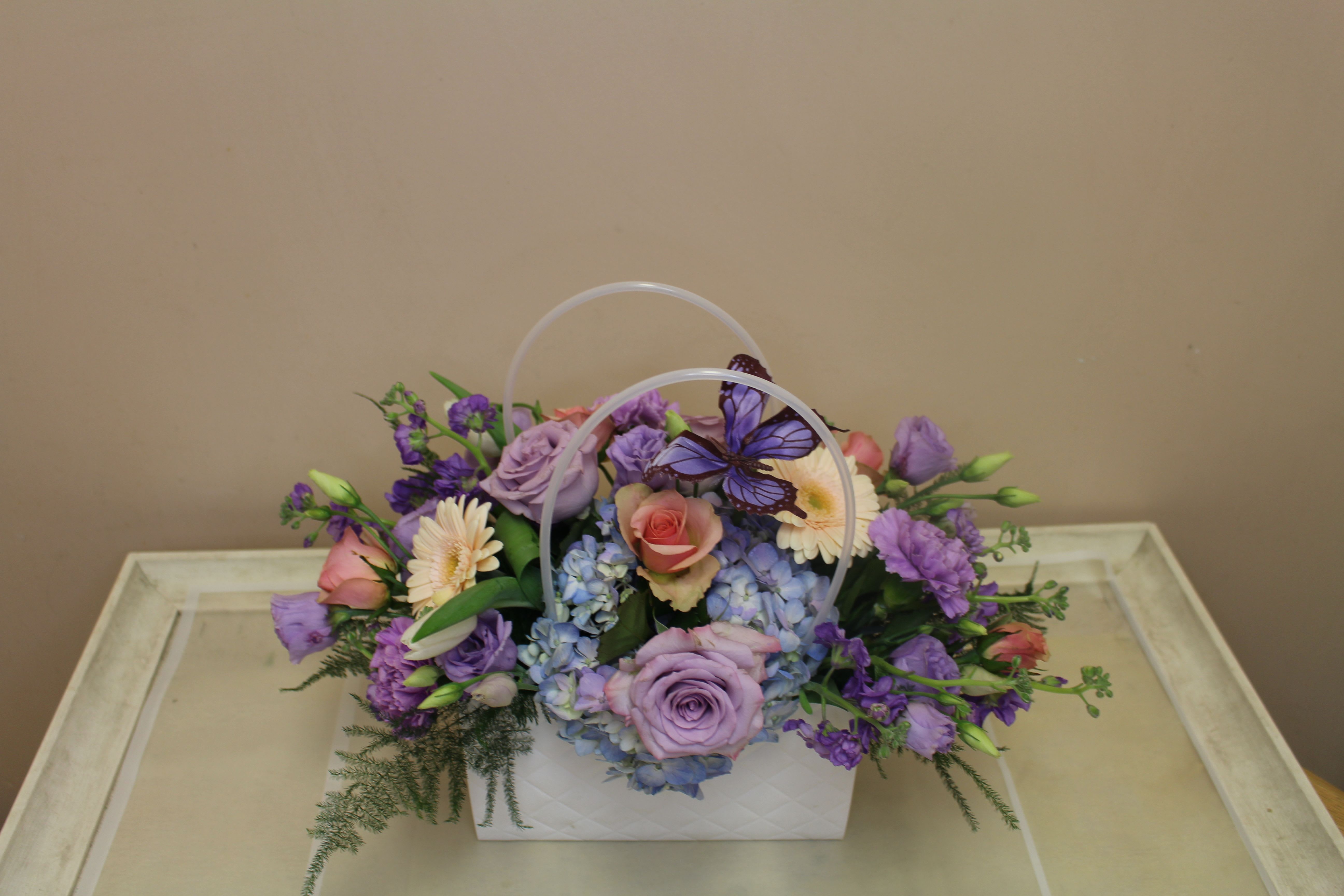 Pocketbook full of Posies Soft - A soft pastel gift arrangement in a decorative white quilted pocketbook, this birthday or spring celebration bouquet mixes lavender and pink roses with pale blue hydrangea and purple lisianthus for a gentle, romantic impression. The decorative purple butterfly perched above the blooms gives the design a whimsical, garden-fresh feel-perfect for birthdays, anniversaries, or a thoughtful thank-you that says you care.  The rounded, abundant composition includes roses, gerbera-style daisies, ruffled carnations, hydrangea clusters, lisianthus and small daisy-like accents arranged with airy trailing greenery. The white pocketbook has an arched clear plastic handle and a quilted look in some versions; sizes present a full, slightly cascading silhouette that brightens any table or bedside.  Made to make the recipient feel truly seen, this arrangement is ideal for someone who loves soft color palettes and a touch of whimsy. Order with standard delivery options; to keep blooms fresh, place in a cool spot away from direct sun and change the water daily if transferring to a vase.