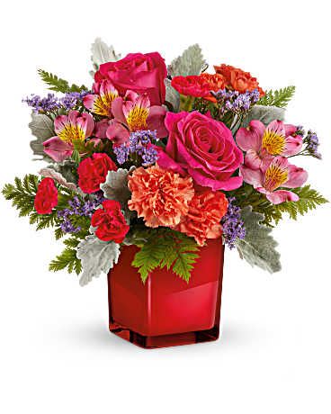 Compact arrangement of pink roses, orange carnations, and mixed blooms in a red cube vase