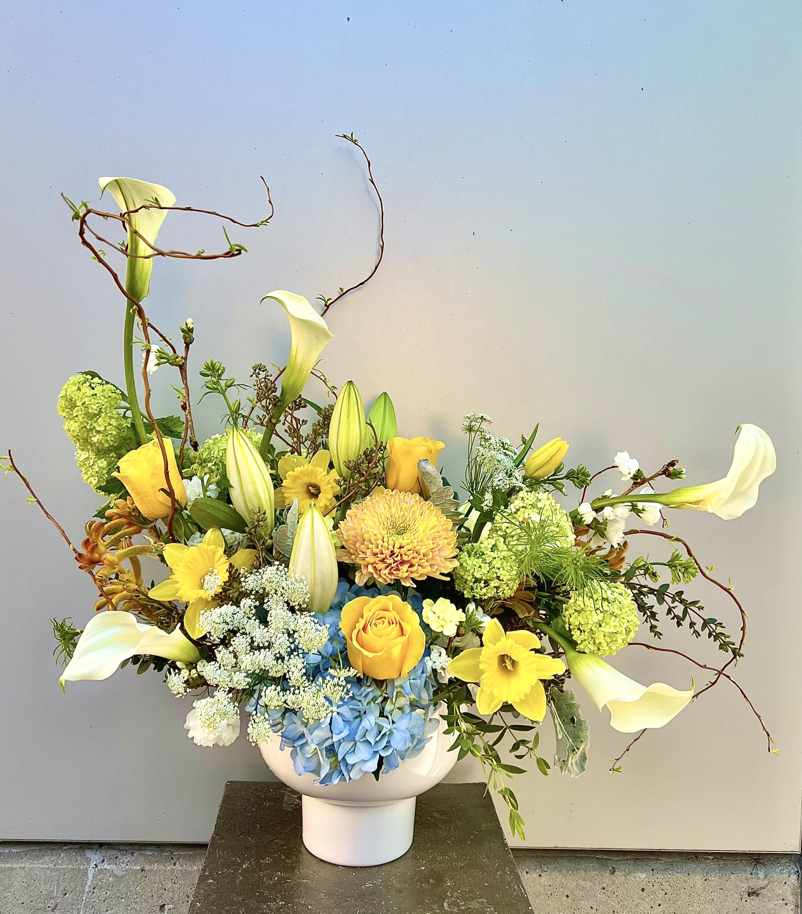 Blooming Bliss - Beautifully crafted floral arrangement in a pedestal vase. Featuring magnificent Calla lilies, delicate yellow roses, Queen Anne's lace, vibrant green viburnum, and a touch of light blue hydrangea, this stunning centerpiece will bring a radiant and sunny ambiance to any space!