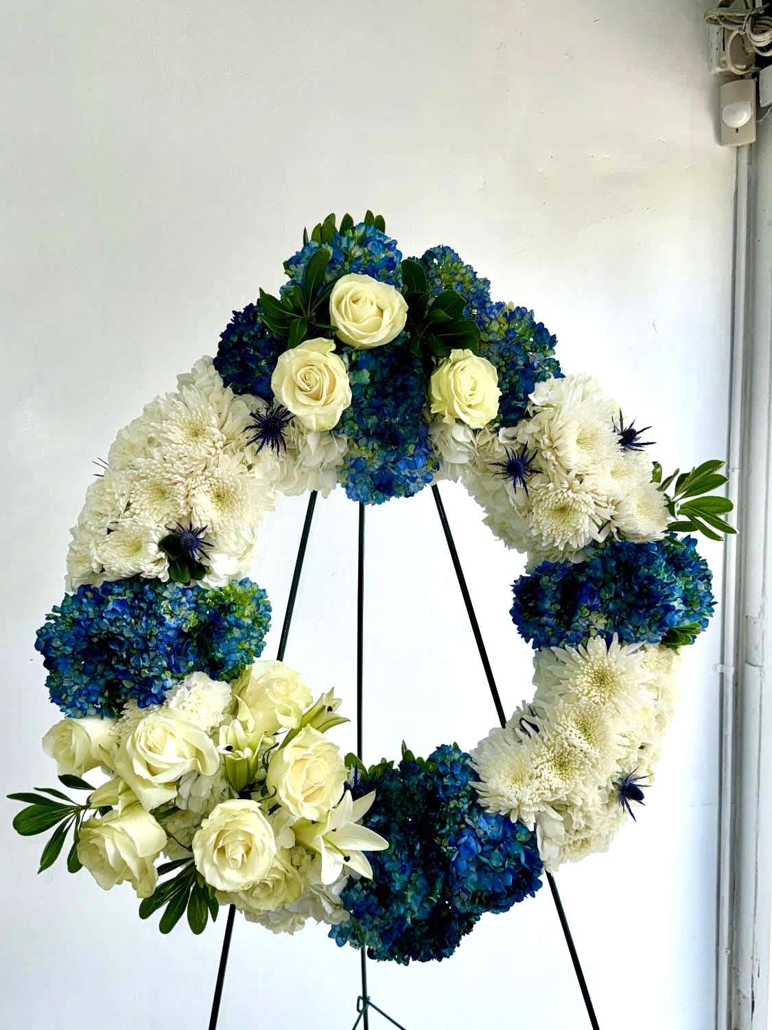 Circular floral wreath with white roses, blue hydrangeas, and white chrysanthemums on a stand